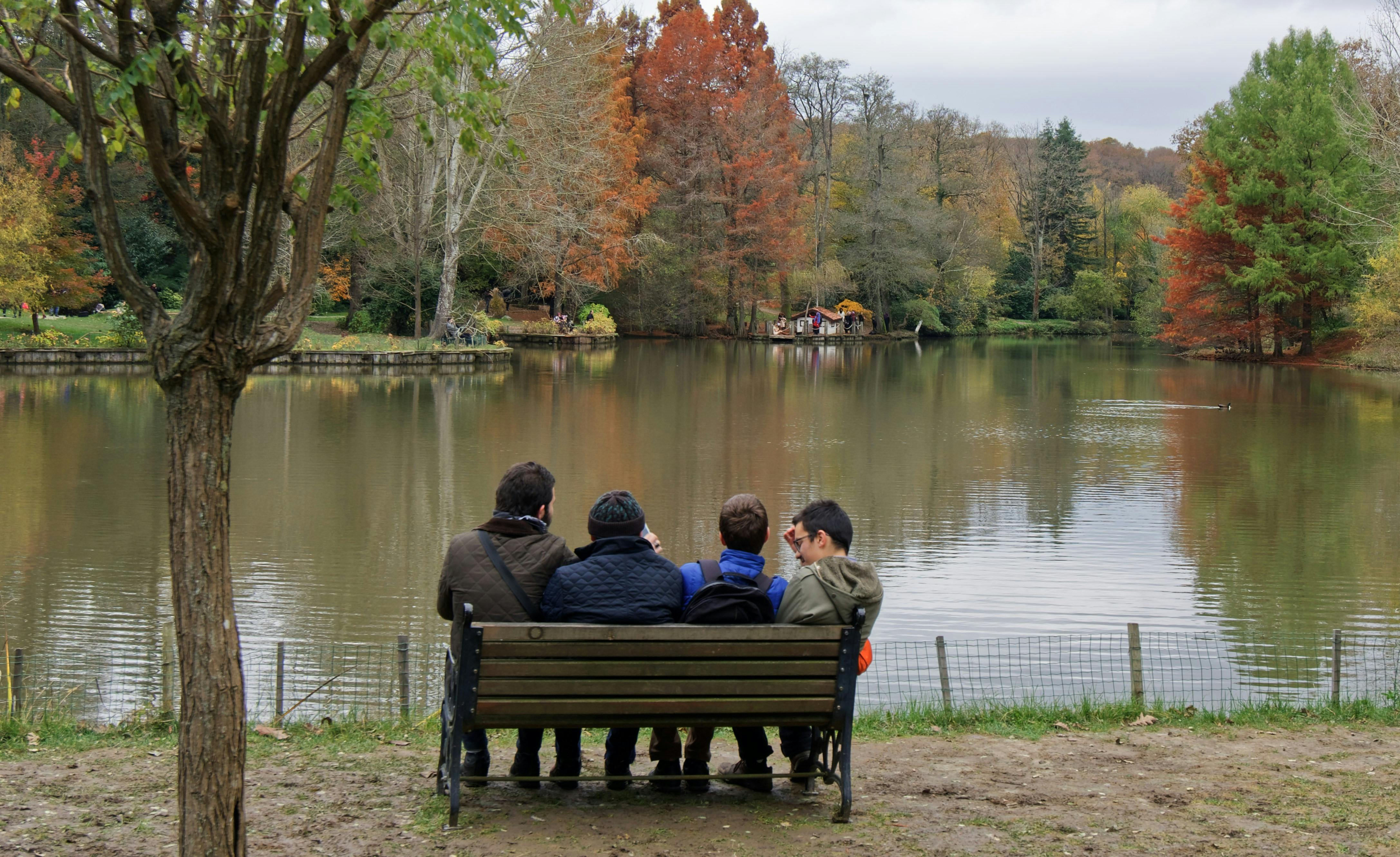 бесплатная Четыре человека сидят на скамейке, наслаждаясь живописным осенним видом у озера в парке. Стоковое фото