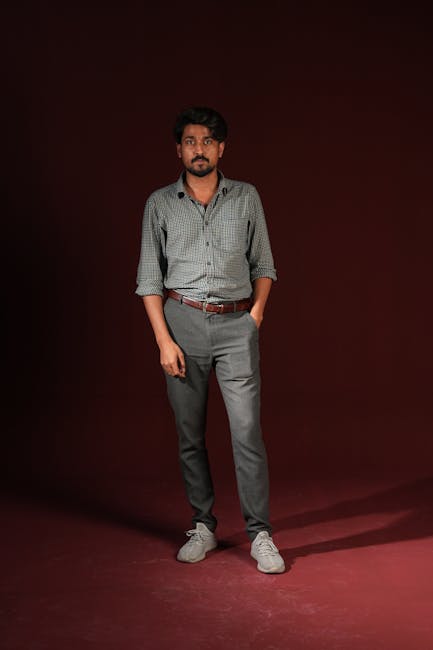Portrait of a young adult man standing against a dark background with hands in pockets, dressed in casual attire.