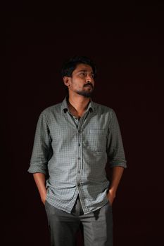 Contemplative man in checkered shirt stands against a dark background, hands in pockets.