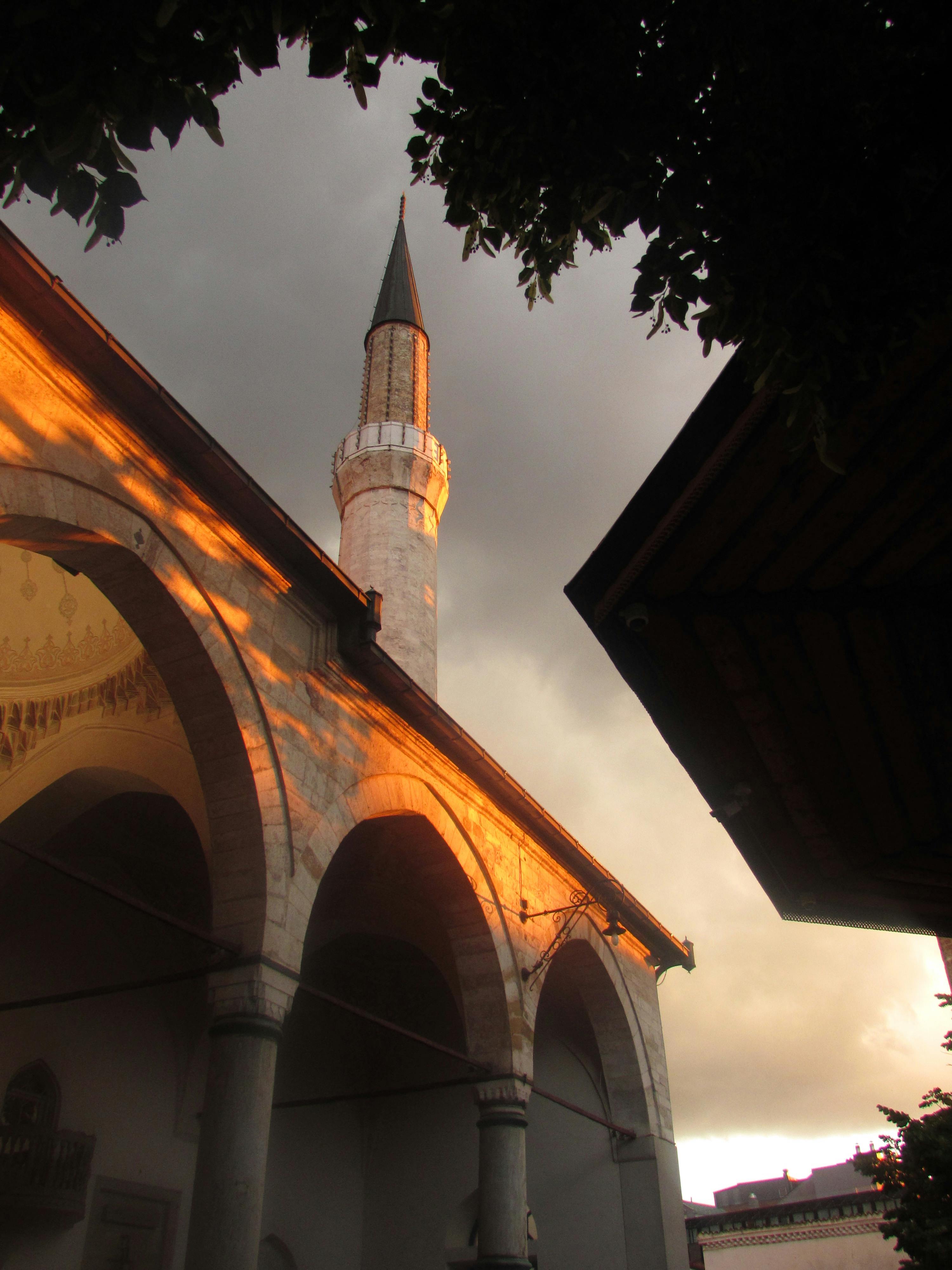 бесплатная Золотистый закат освещает мечеть Гази Хусревбега в Сараево, создавая потрясающий контраст с темными облаками. Стоковое фото