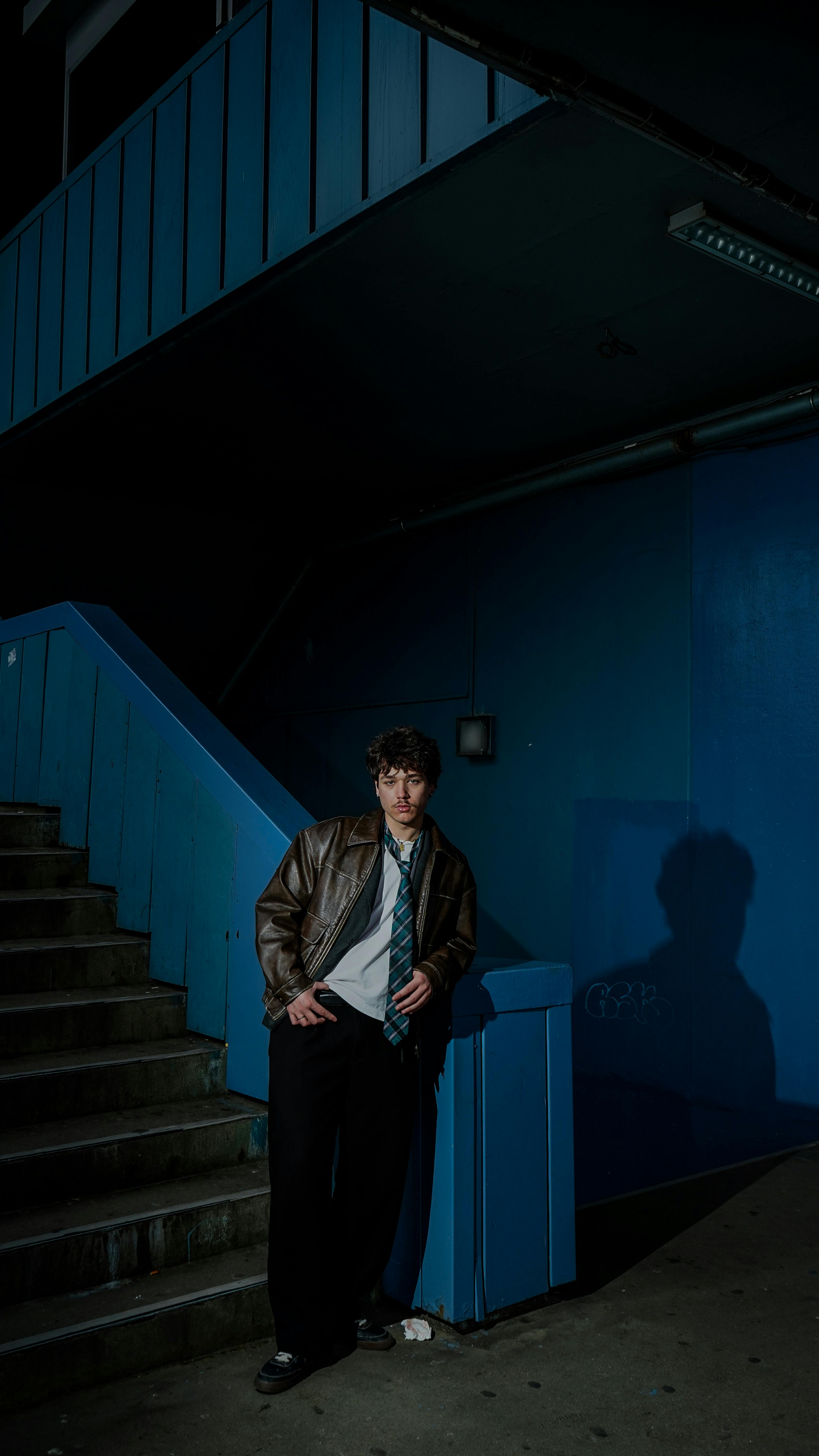 無料 革のジャケットを着た若い成人が青い壁に寄りかかり、都会の階段、カジュアルなポーズ、夕方の設定。 写真素材