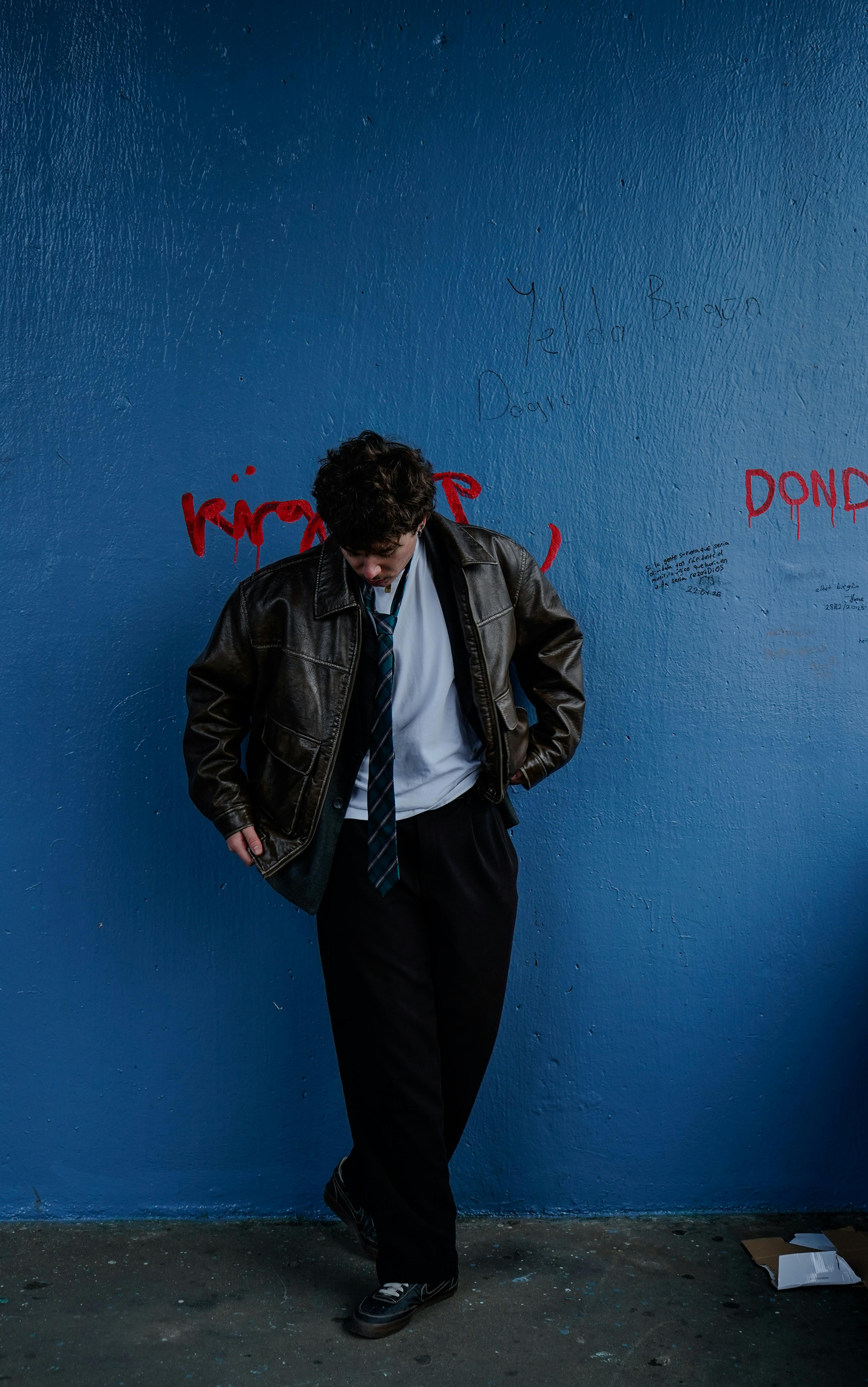 бесплатная Мужчина в кожаной куртке задумчиво стоит на фоне стены, разрисованной синими граффити. Атмосфера городской моды. Стоковое фото