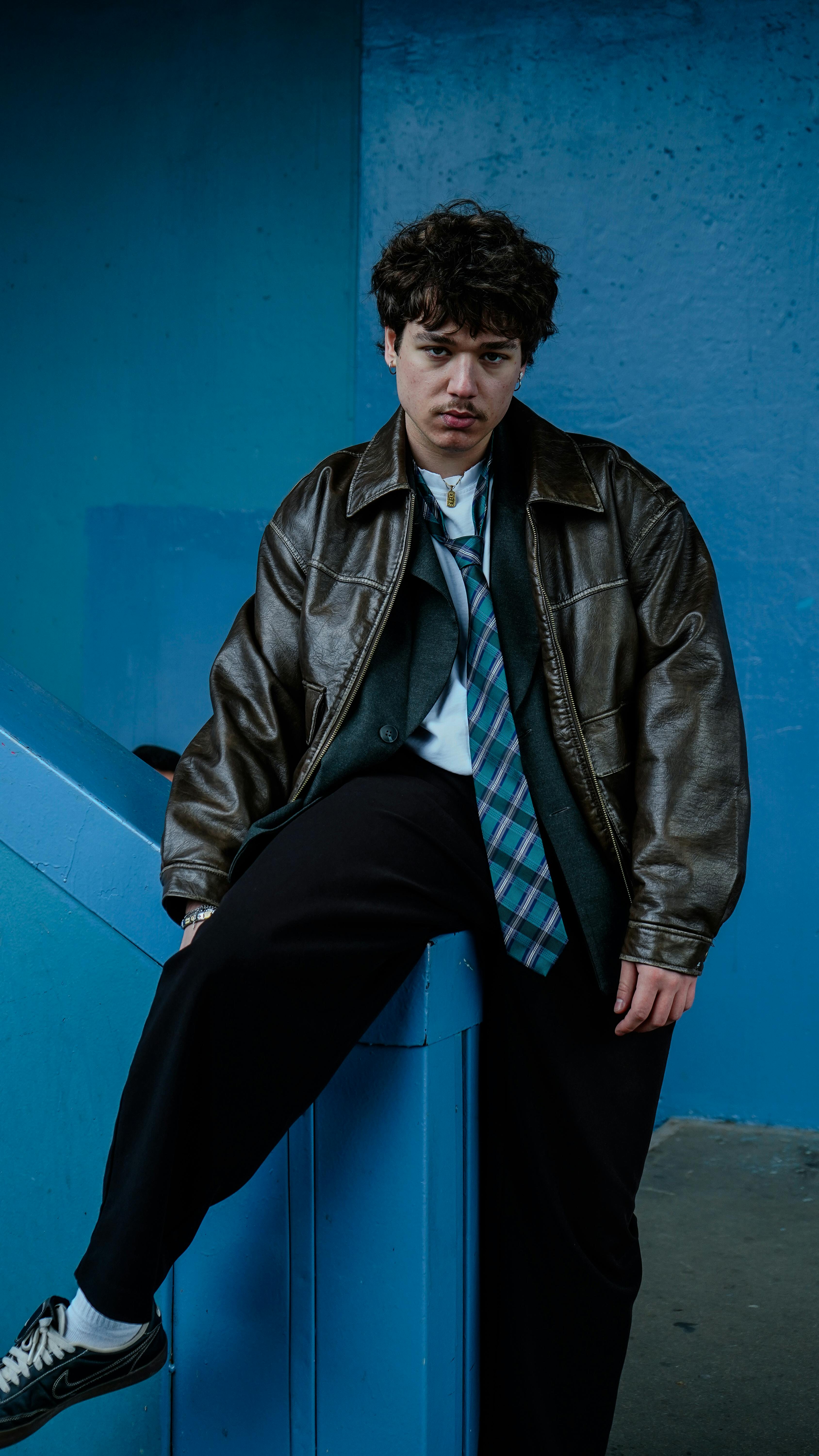 Free Young man in a leather jacket leaning on a blue wall, daytime urban setting. Stock Photo