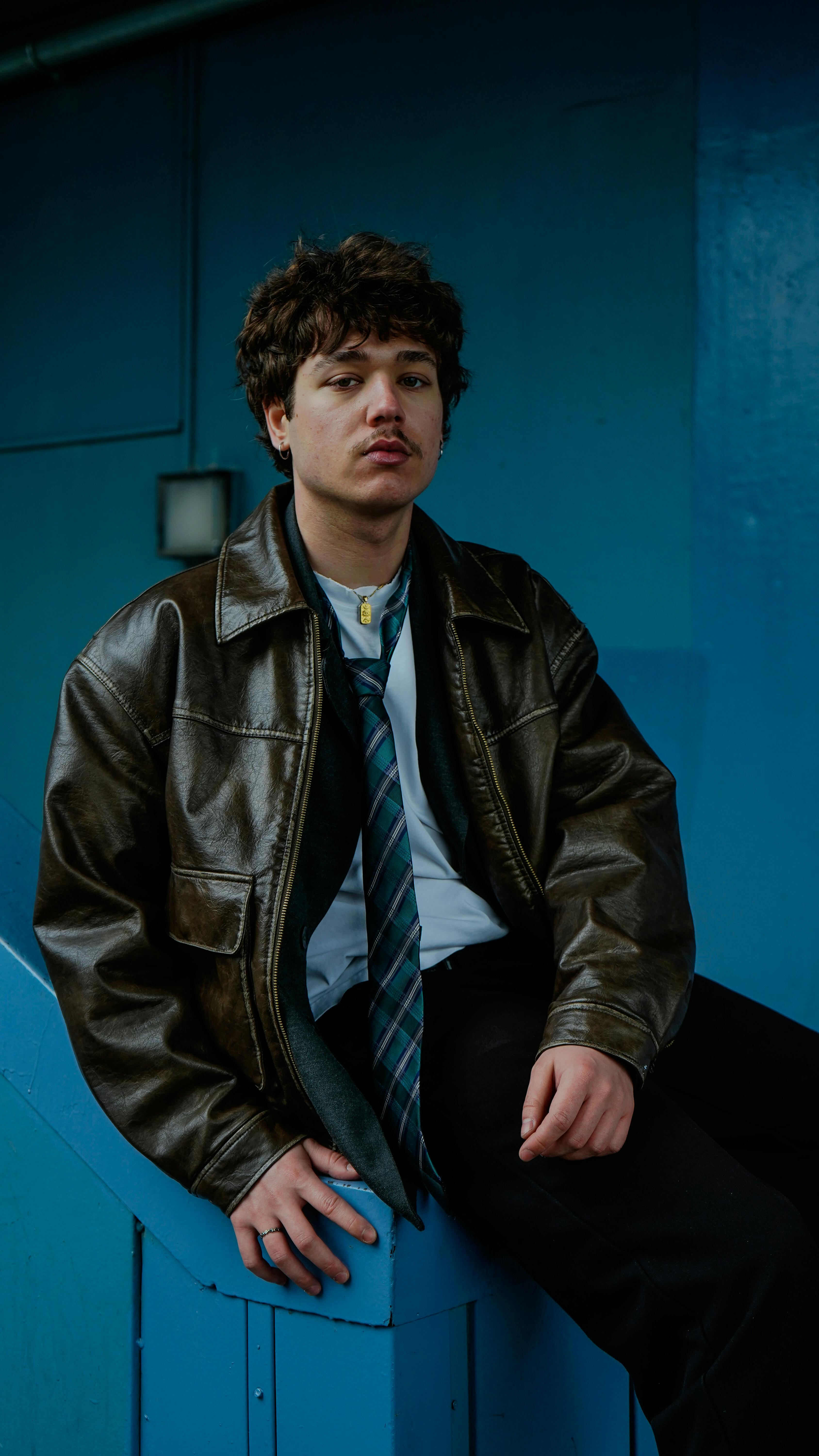 Gratis Seorang pria muda mengenakan jaket kulit dan dasi berpose di depan dinding biru yang cerah. Foto Stok