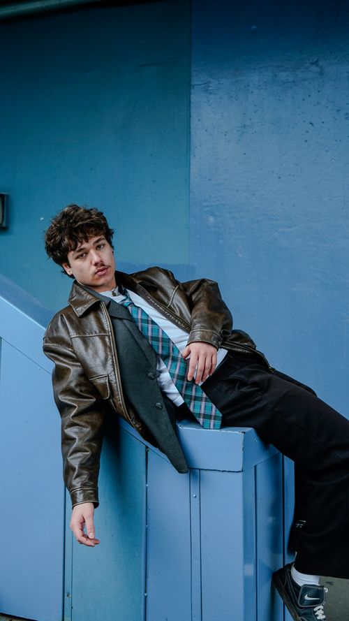Free Young man in leather jacket leaning on blue background, urban style portrait. Stock Photo