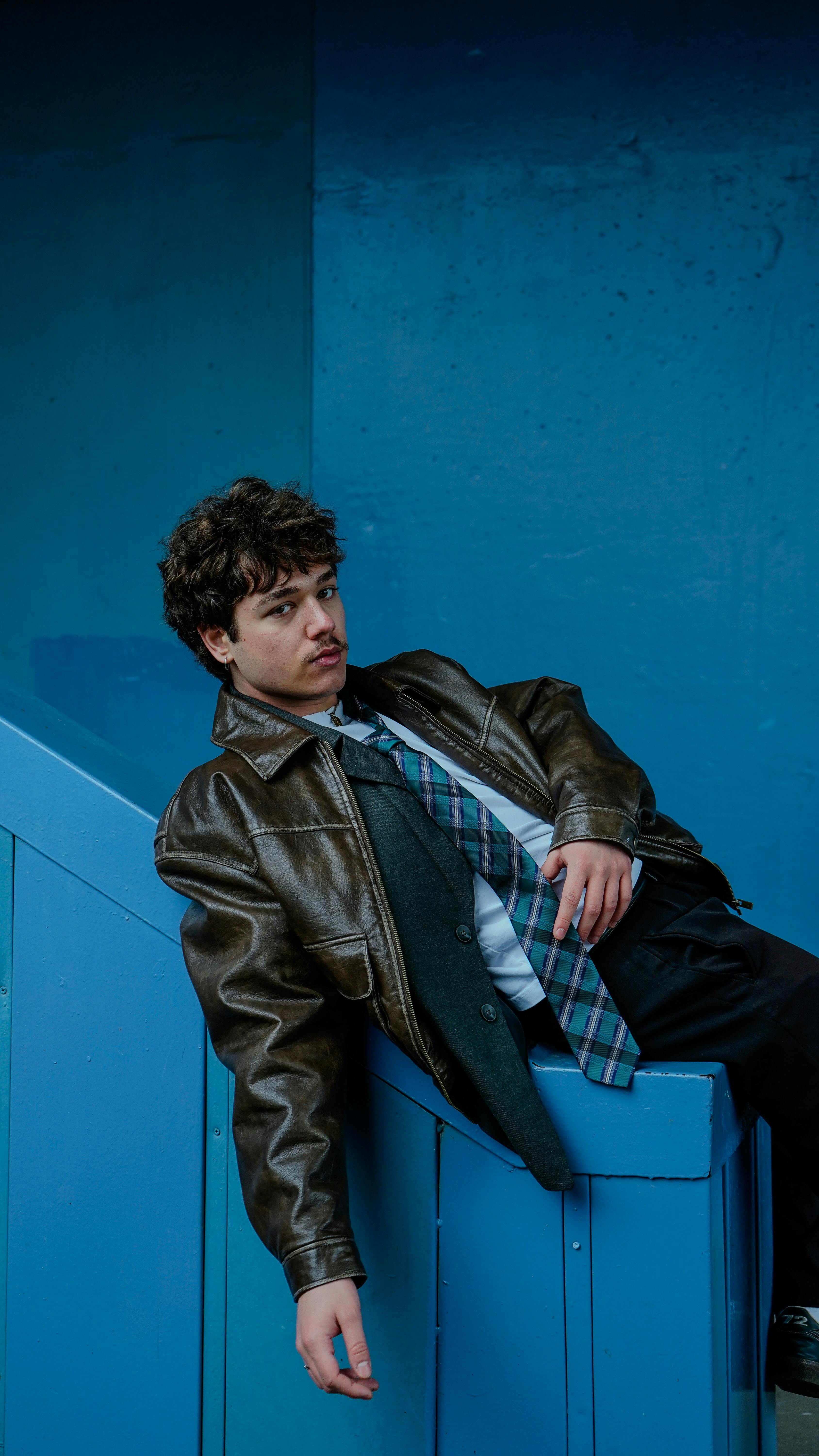 無料 革のジャケットとネクタイを身に着けた若い男性が、鮮やかな青い壁を背景にスタイリッシュにポーズをとっています。 写真素材