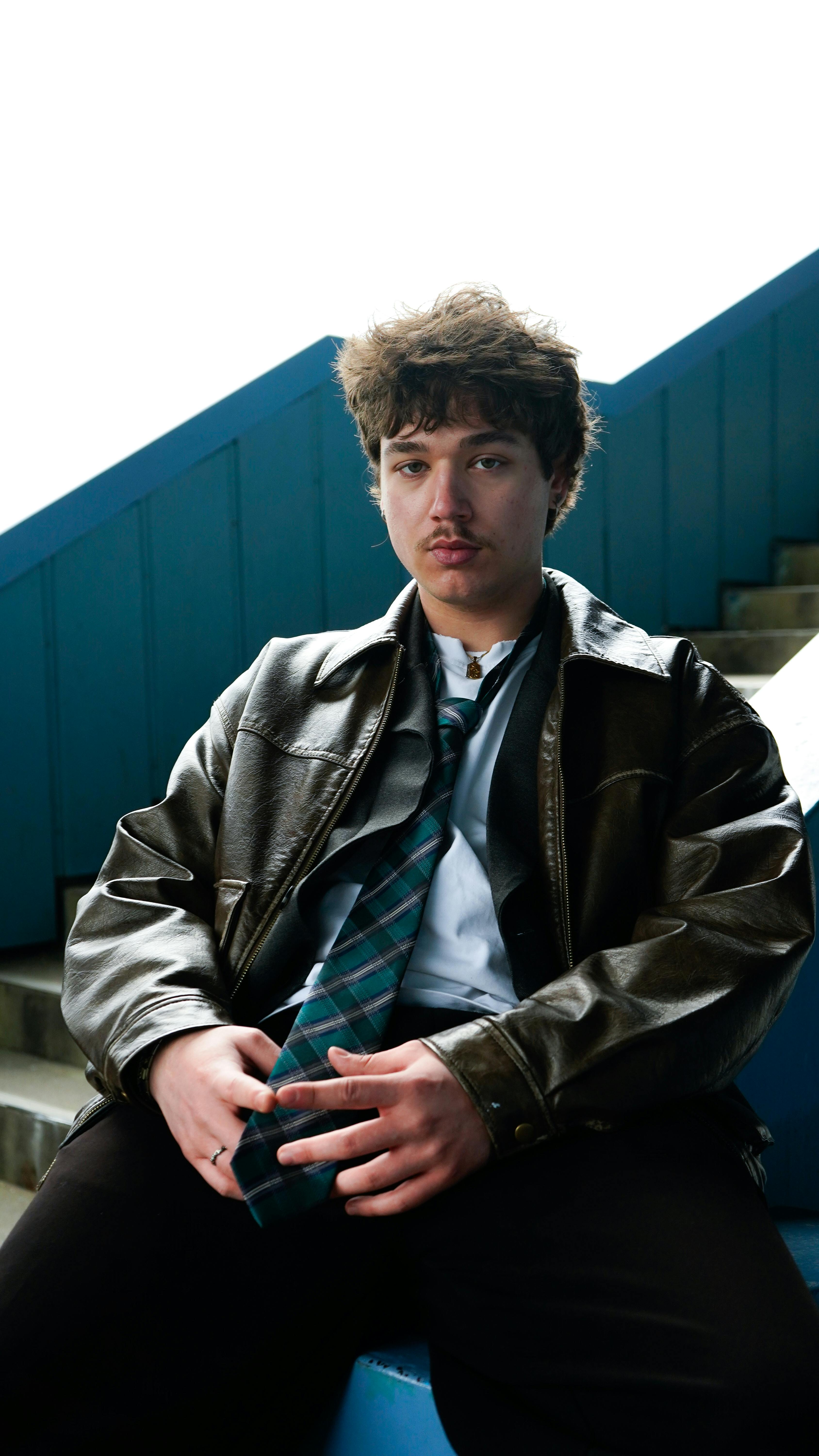 Free Young adult male in a leather jacket and tie sitting on blue stairs outdoors. Stock Photo