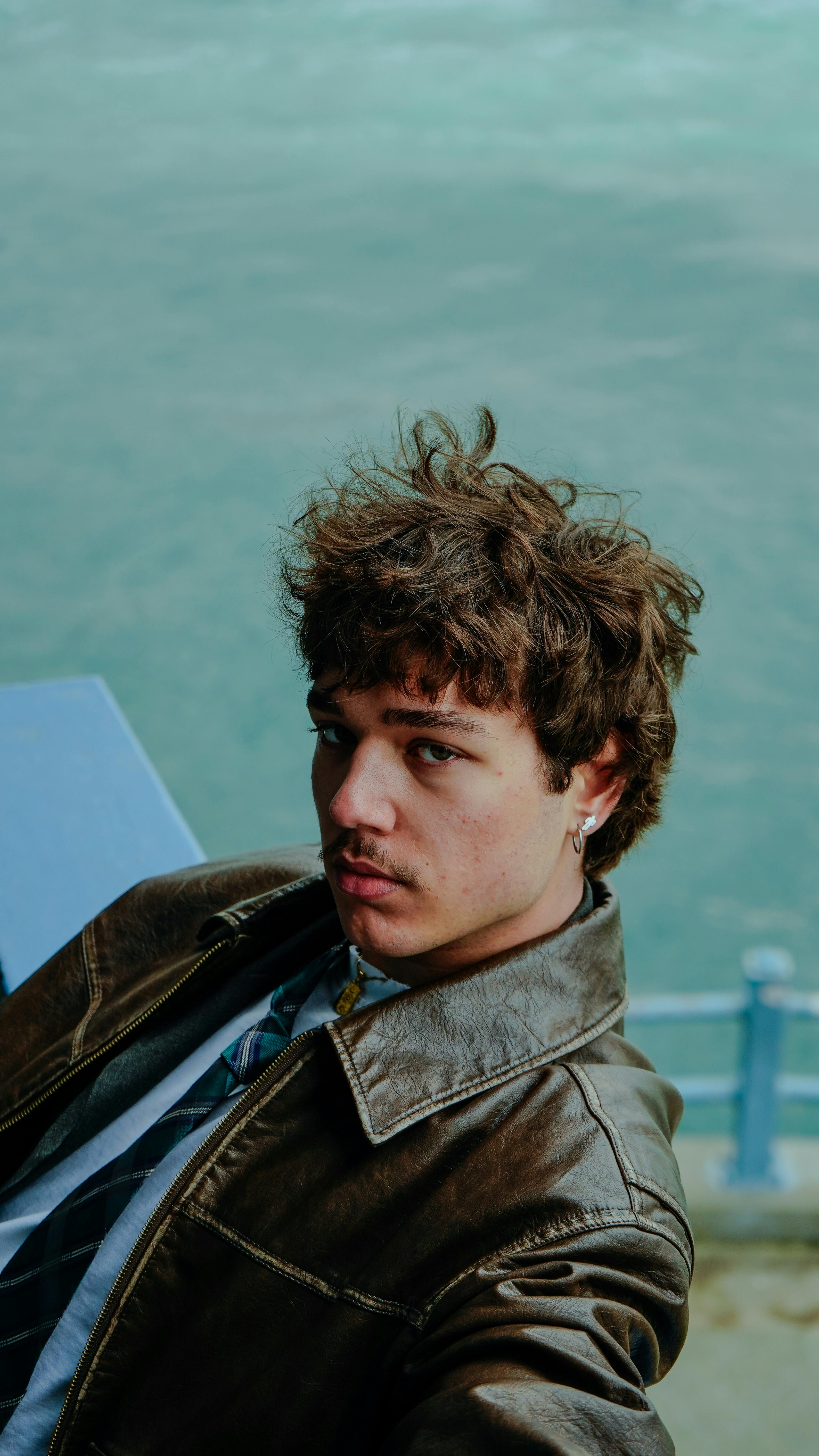 Free Portrait of a young man with curly hair in a leather jacket by a seaside view. Stock Photo