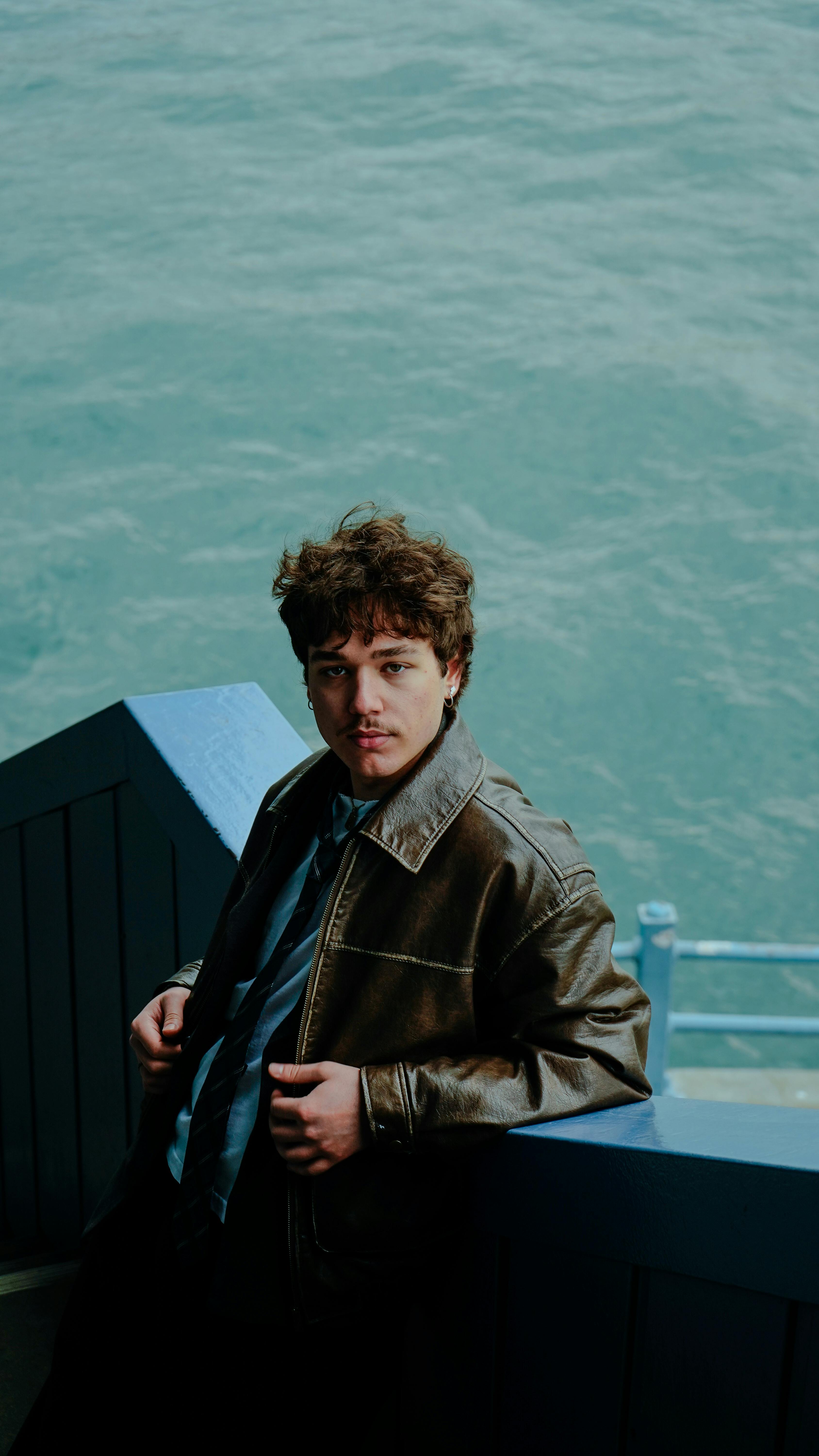 Ücretsiz Soğuk bir günde, deri ceket giymiş genç bir adam su kenarında rahat bir poz veriyor. Stok Fotoğraflar