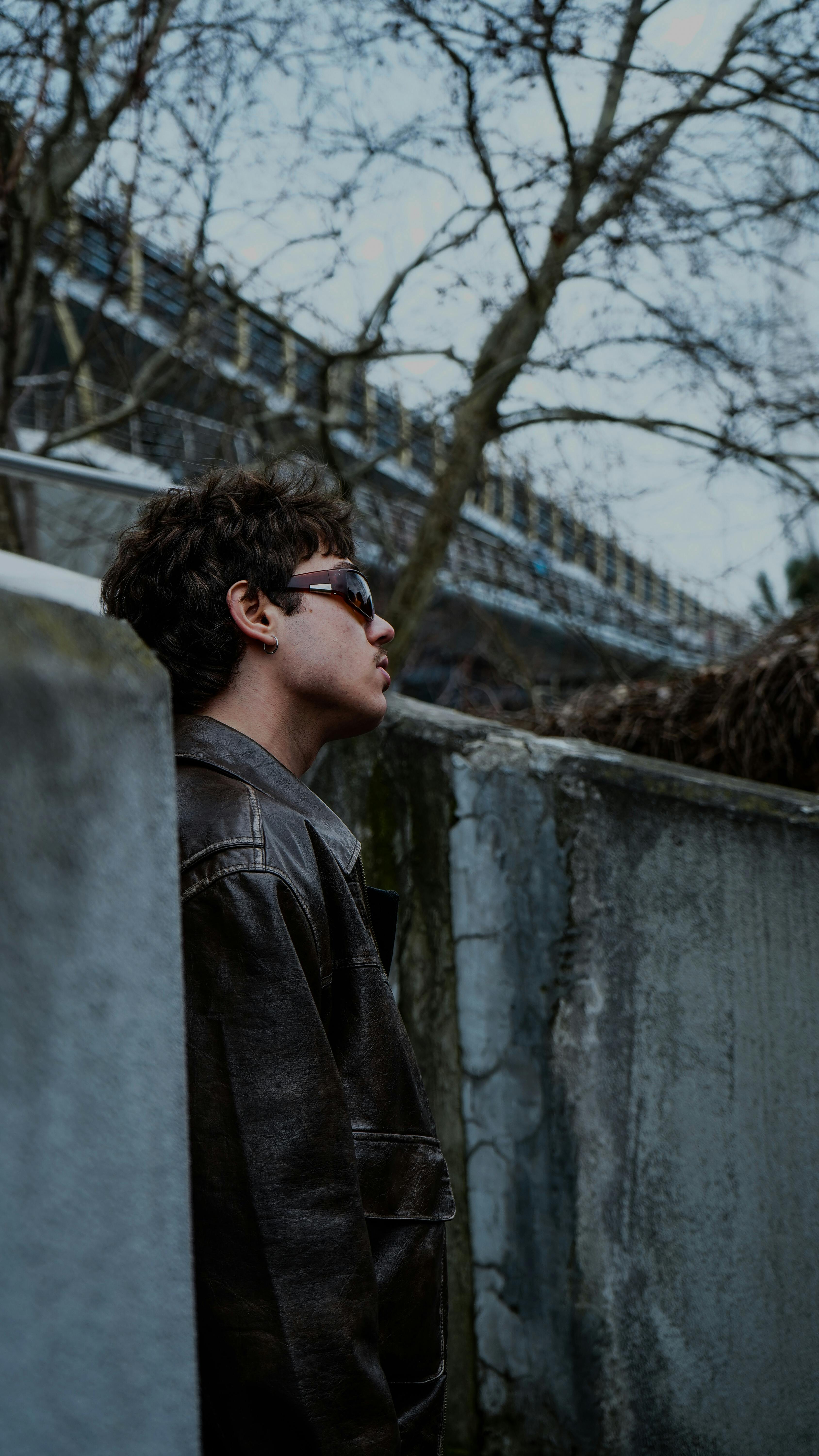 Free Young man in sunglasses leans on a wall, exuding a cool vibe on an overcast day. Stock Photo