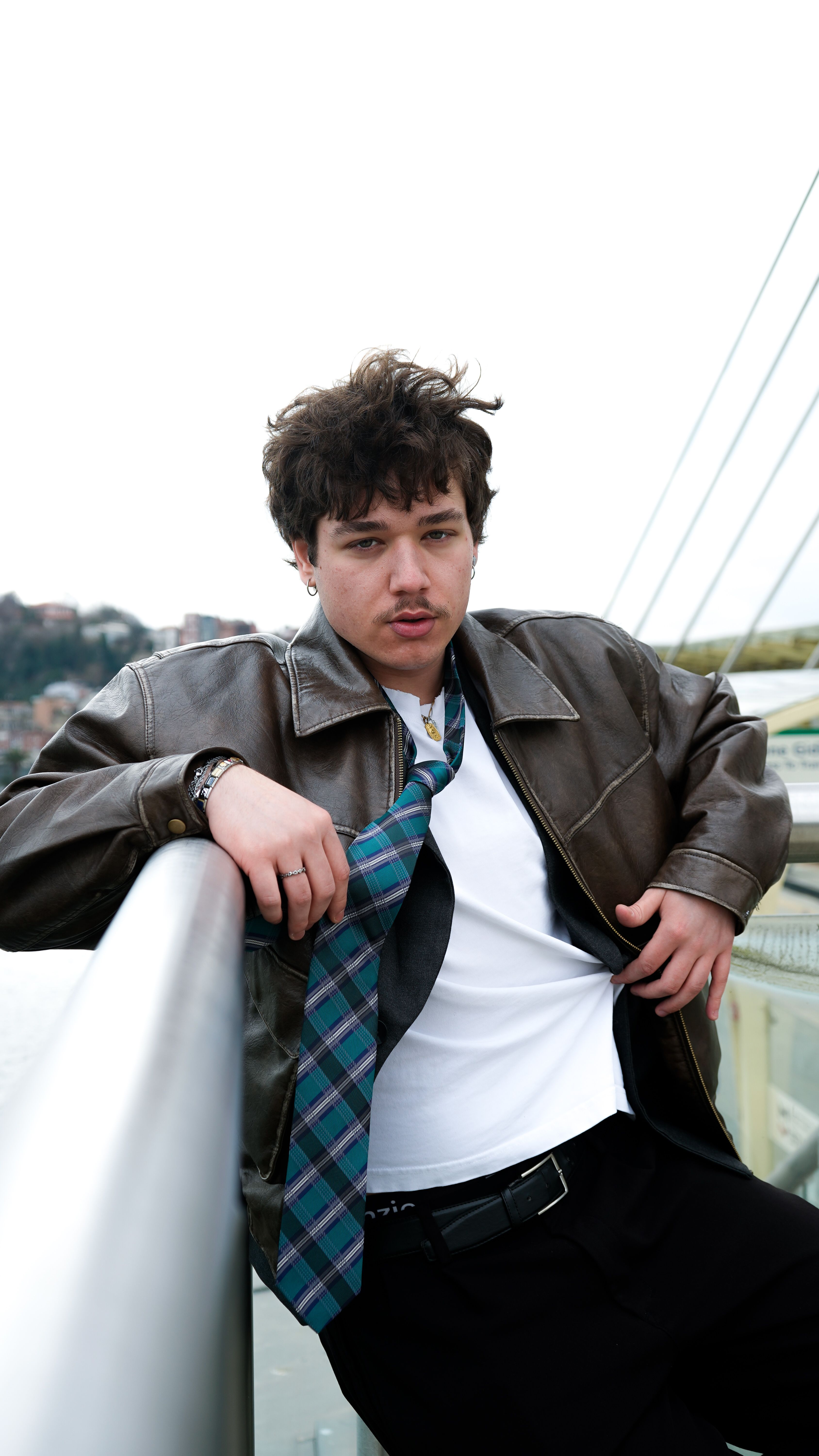Casual portrait of a young man leaning on a bridge railing with city skyline in background.