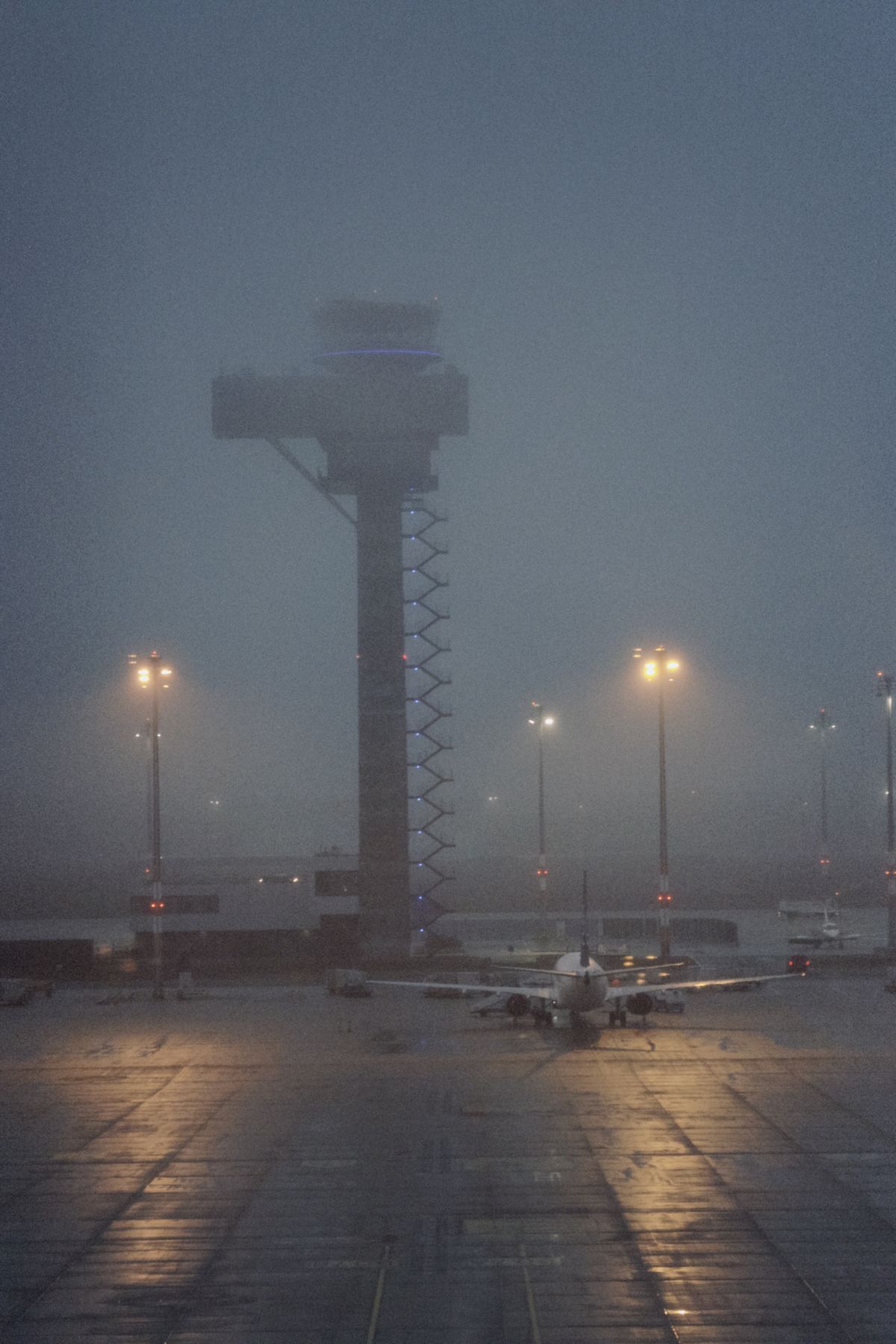 A misty airport apron at dusk with a control tower and an aircraft barely visible through the fog