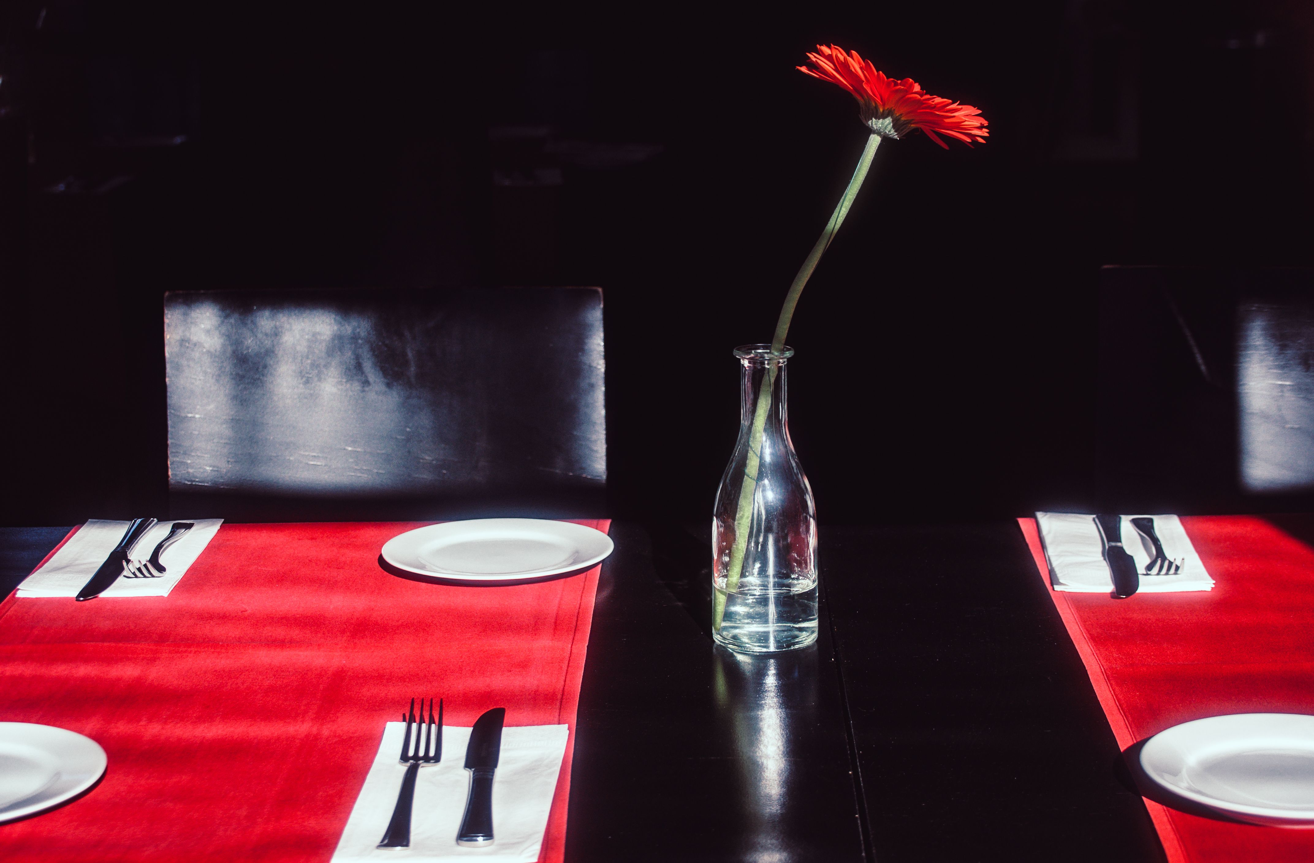 A stylish restaurant table setting featuring a single red Gerbera in a vase on a red tablecloth.