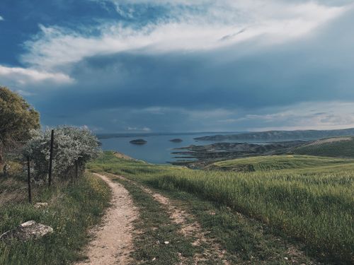 Free A serene dirt path through lush fields leading to a picturesque lake under a dramatic sky. Stock Photo