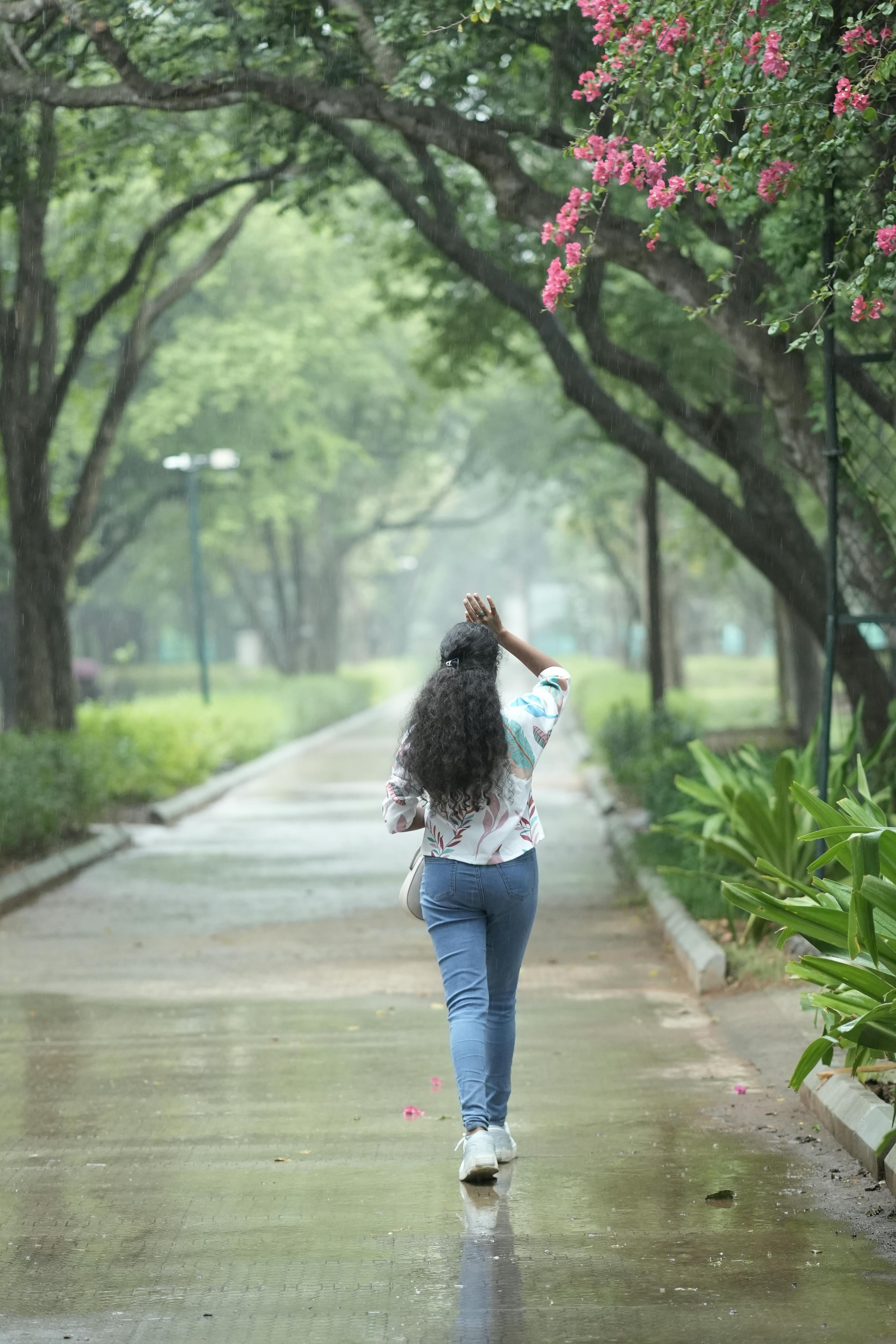 無料 雨の日に、鮮やかな葉とピンク色の花に囲まれた緑豊かな公園を散歩する女性。 写真素材