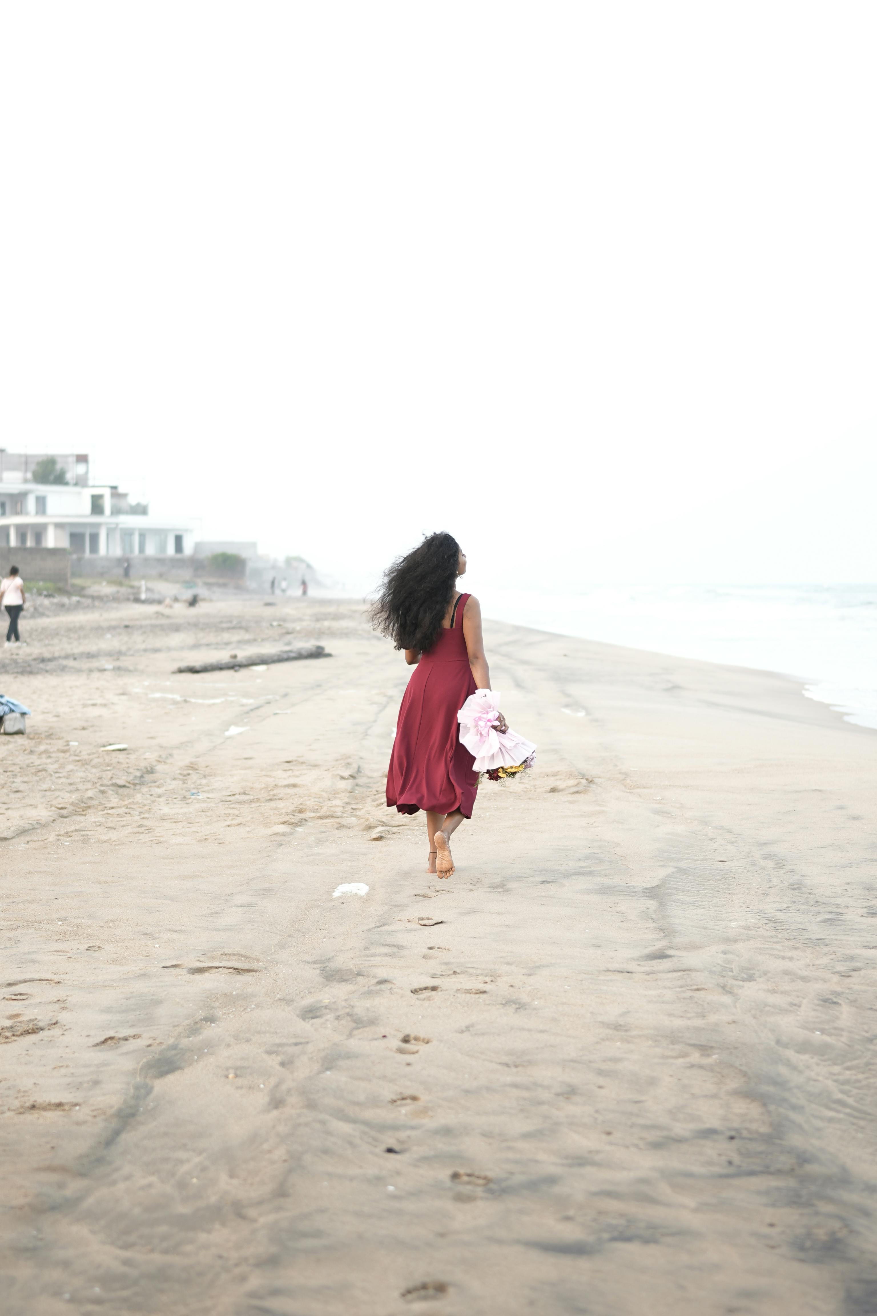 無料 赤いドレスを着た女性が花を持って静かな海岸を歩いており、平和な孤独を表しています。 写真素材