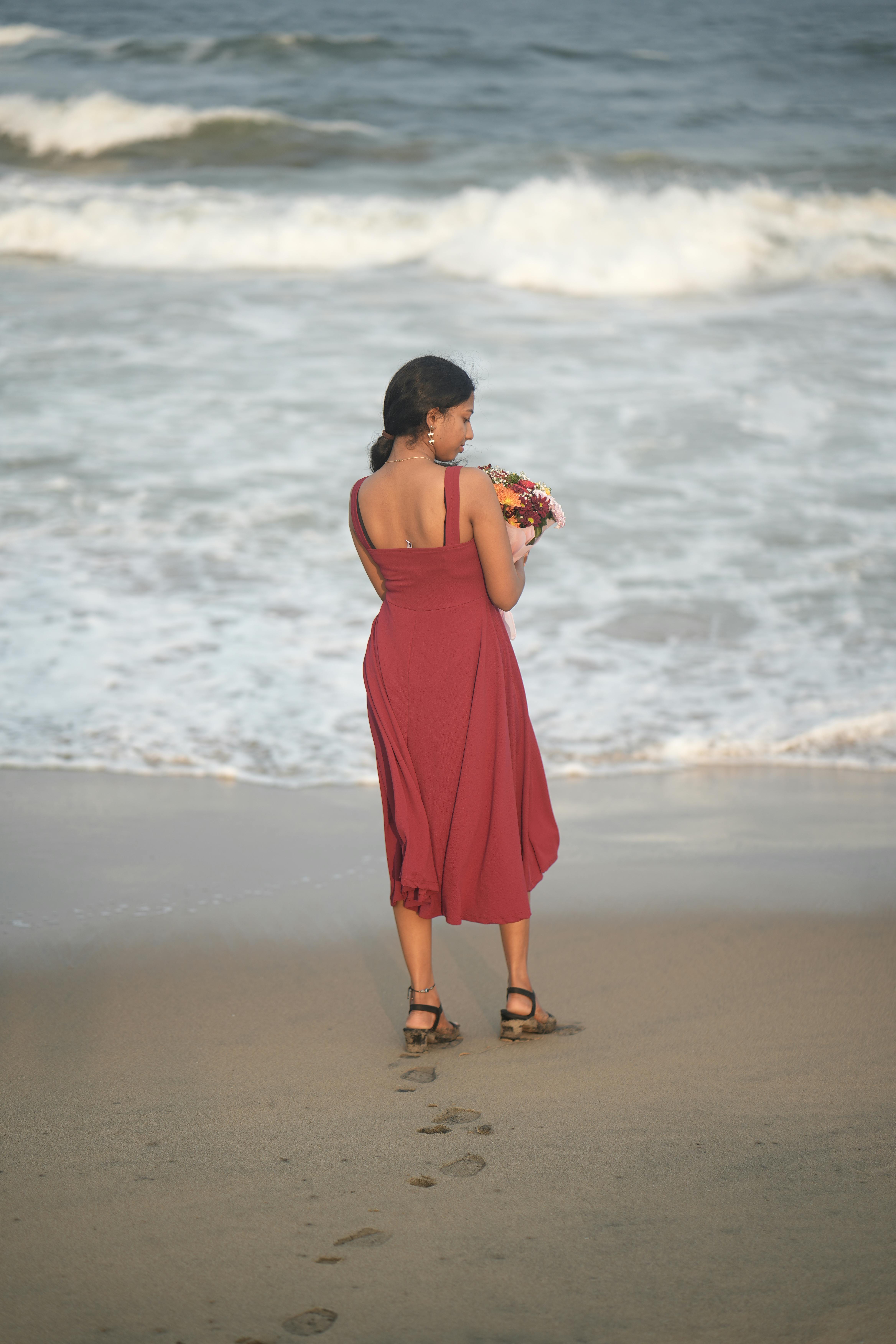 бесплатная Женщина в красном платье с букетом в руках на спокойном пляже, окруженном океанскими волнами. Стоковое фото
