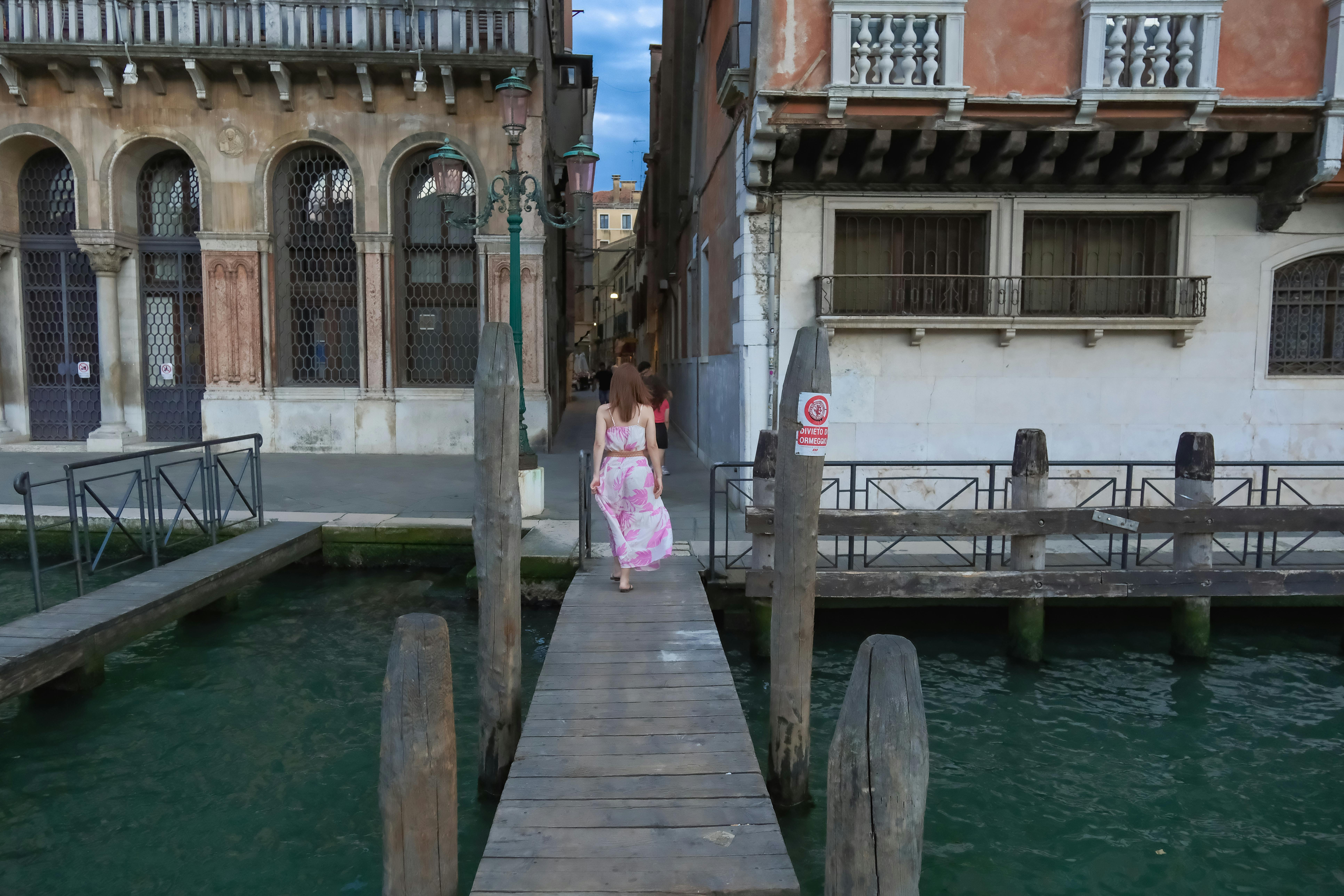 無料 花柄のドレスを着た女性がベネチアの運河沿いの木製の桟橋に沿って歩いています。 写真素材
