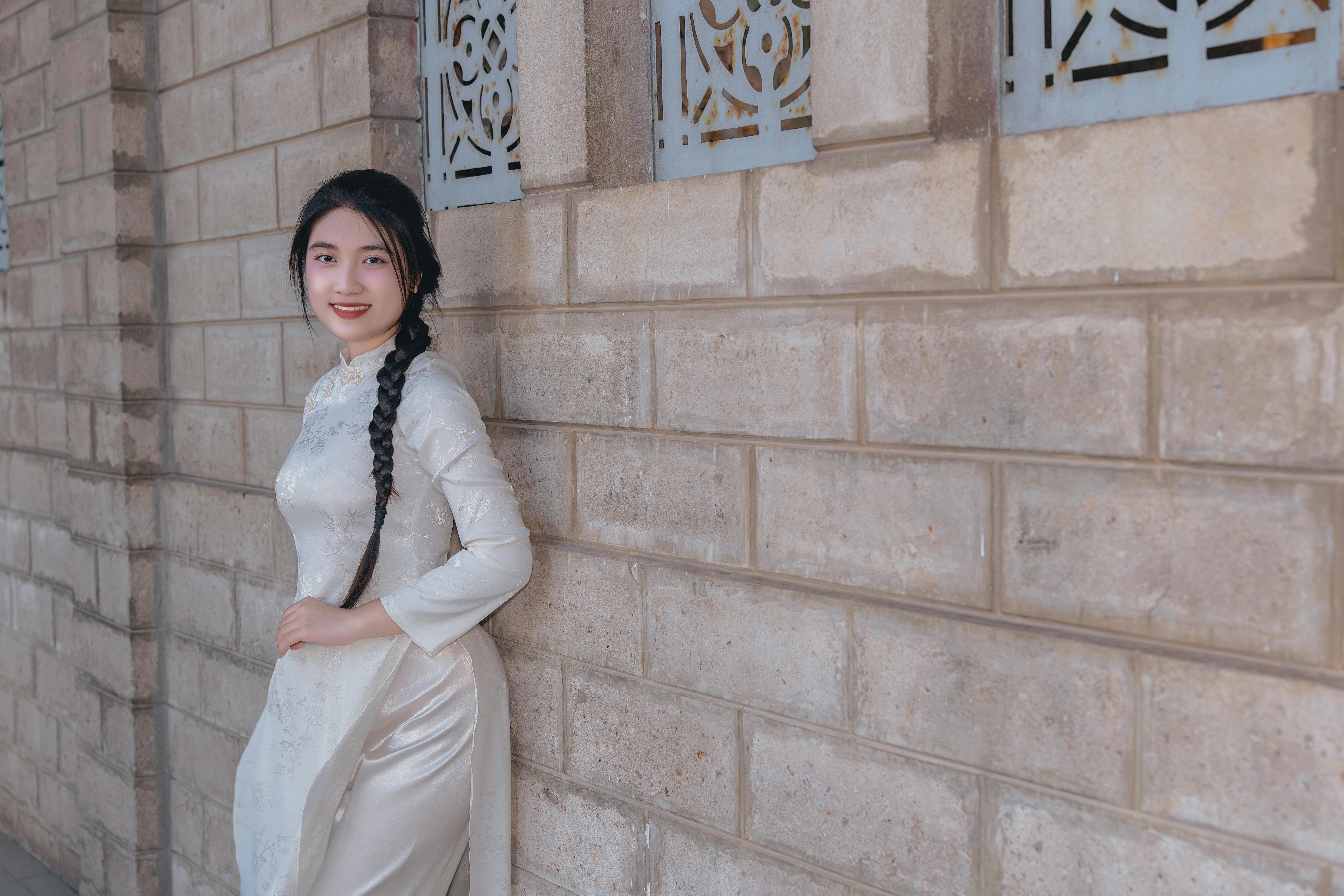 Gratis Potret seorang wanita muda mengenakan gaun tradisional putih dengan latar belakang dinding bata di luar ruangan. Foto Stok