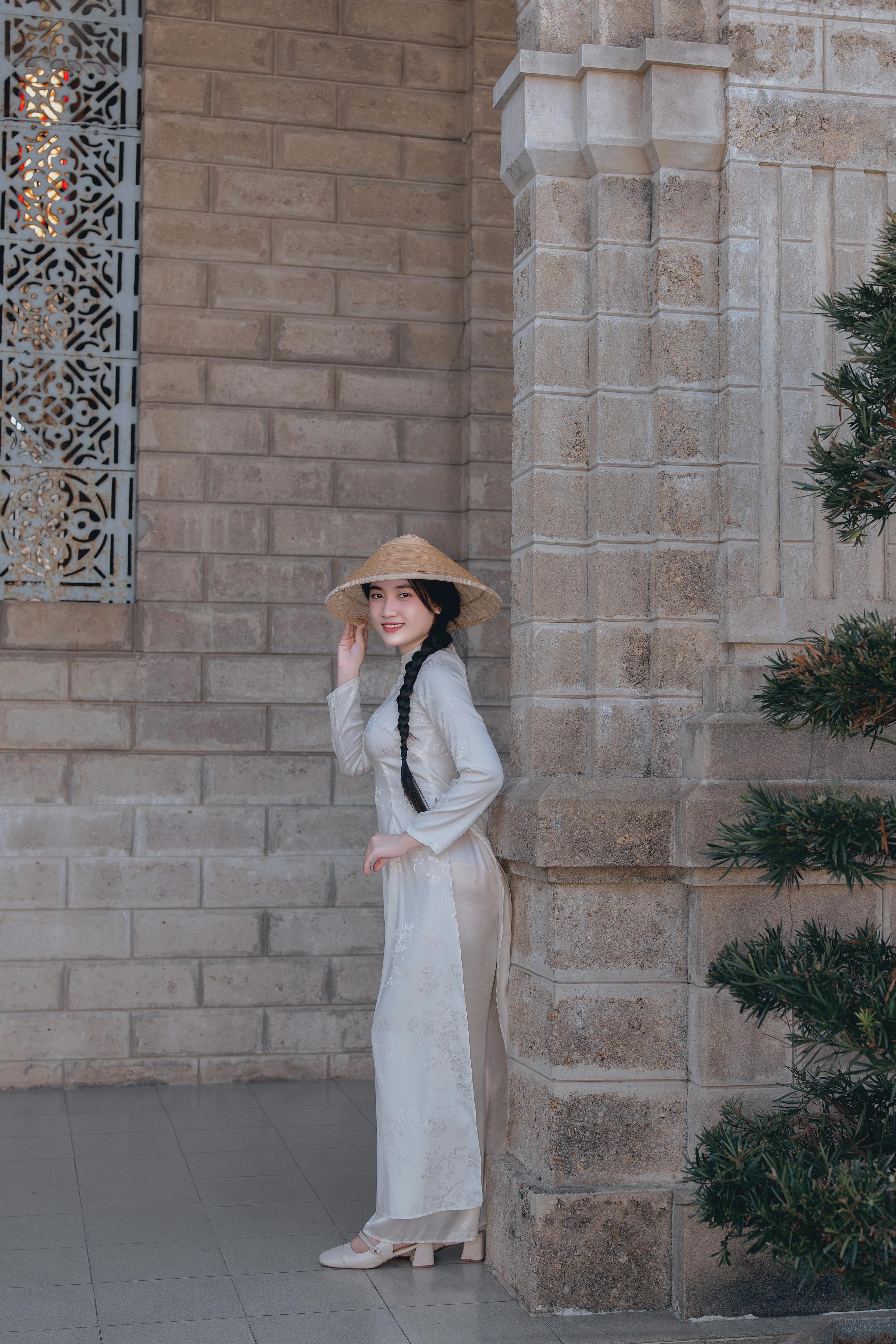 無料 歴史的建築物の横に立つ、アオザイと円錐形の帽子をかぶった優雅な女性。文化的な優雅さを体現しています。 写真素材