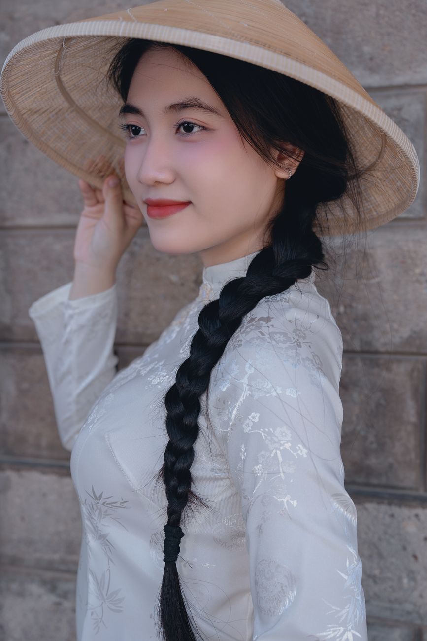 Portrait of a woman in traditional Vietnamese attire and conical hat against a stone wall.