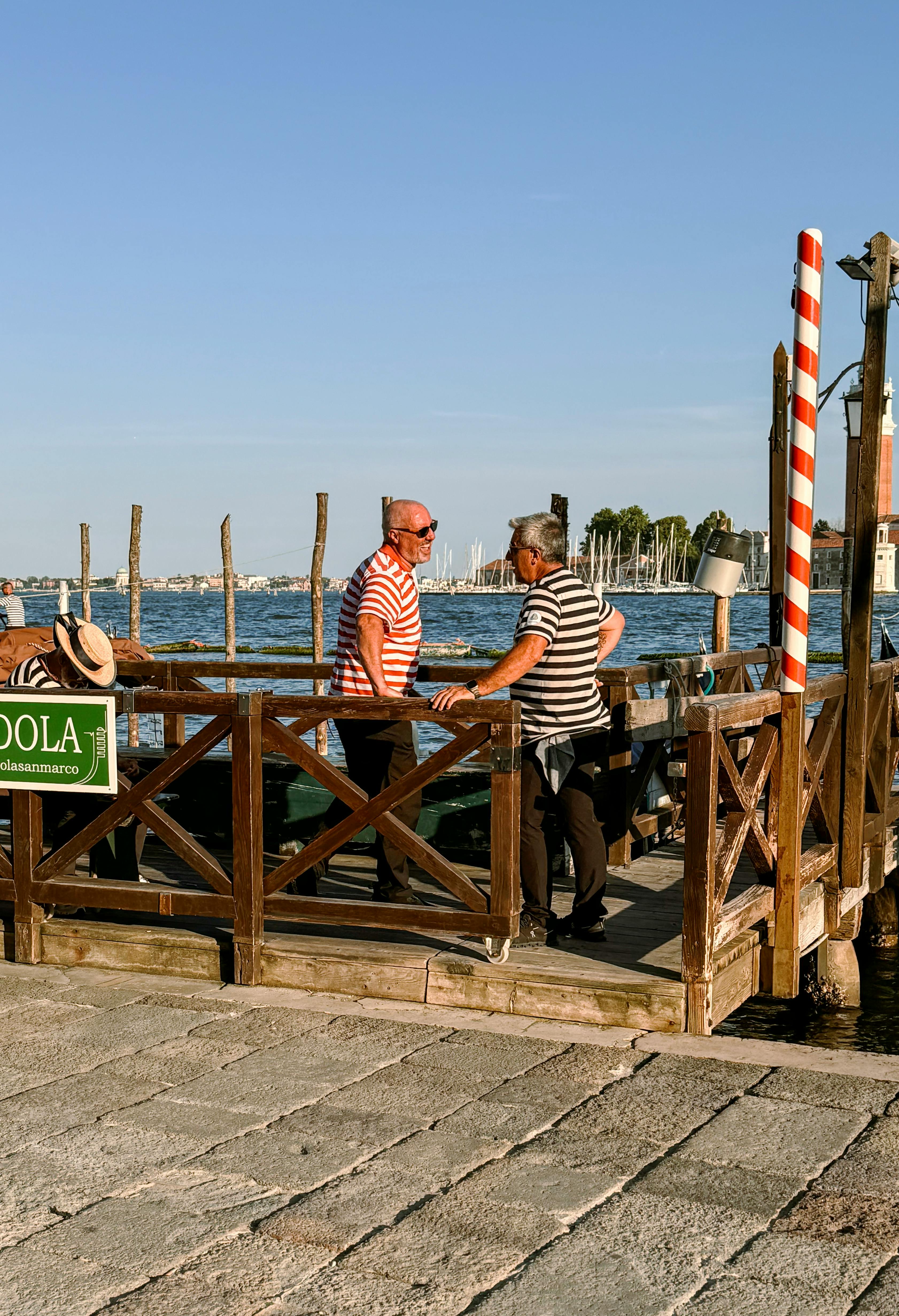 無料 穏やかな海の景色と海をイメージした装飾が施されたイタリアのベニスの陽光降り注ぐ桟橋で、2人の男性が会話を交わしています。 写真素材