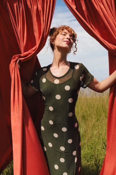 A woman in a polka dot dress enjoys the sunlight while standing between vibrant red curtains.