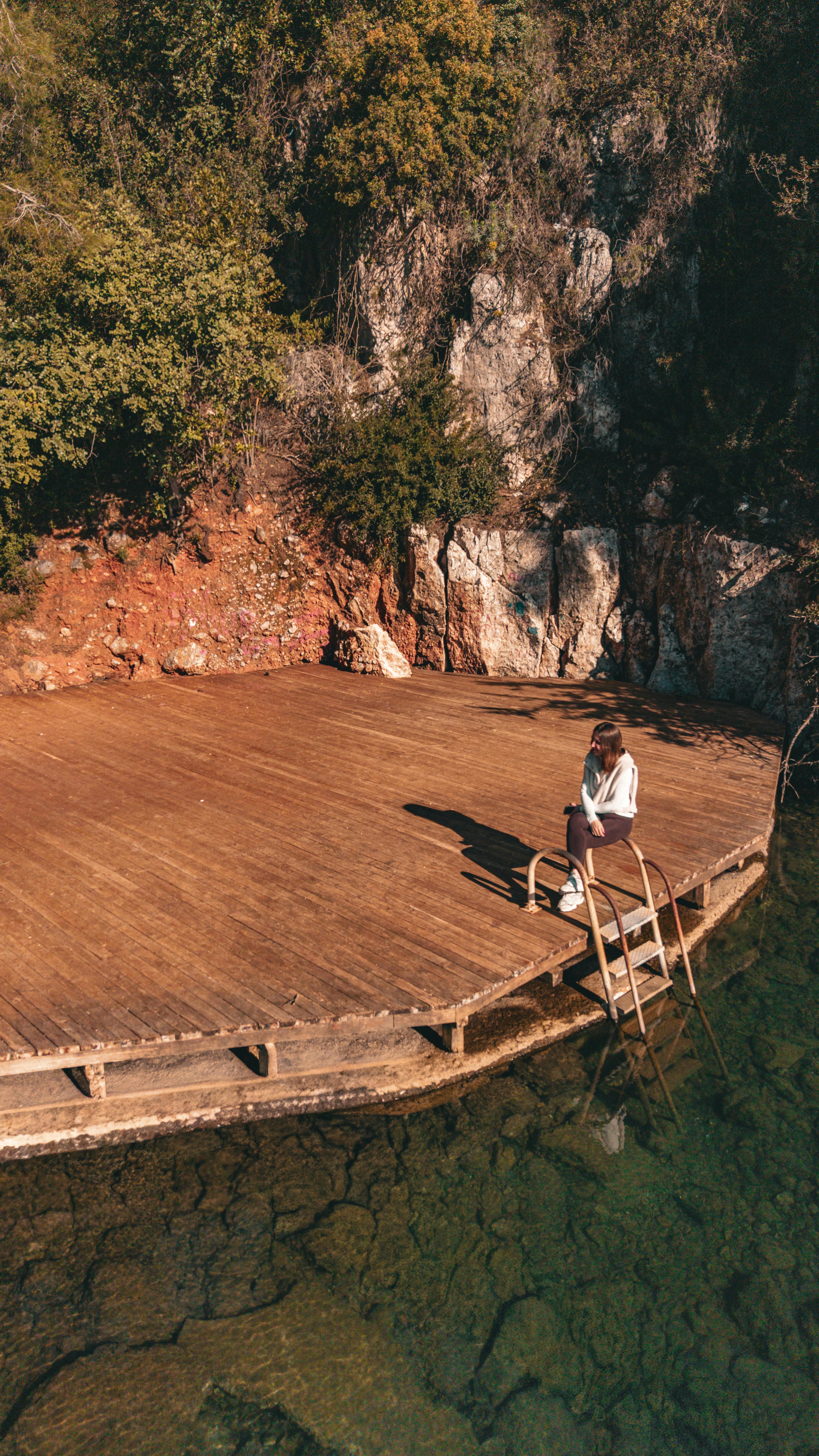Free A woman relaxes on a wooden deck overlooking a tranquil lake with rocky and forested surroundings. Stock Photo