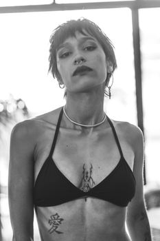 Black and white portrait of a stylish tattooed woman wearing a bikini indoors.