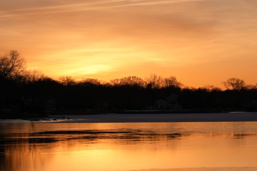 Beautiful winter sunset casting golden hues over a calm lake with silhouetted trees.