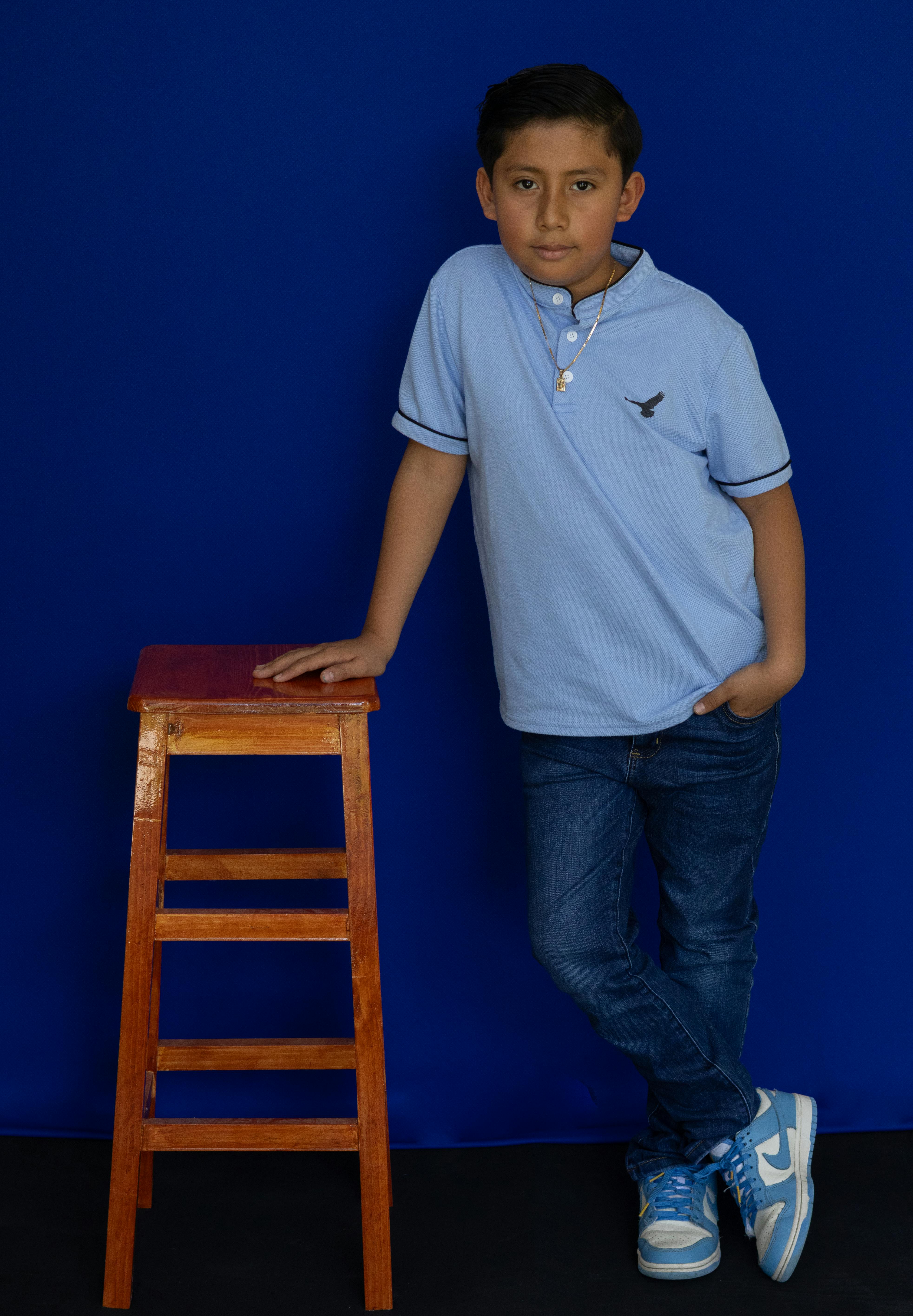 Free Young boy in casual attire leaning against a wooden stool, blue backdrop. Stock Photo