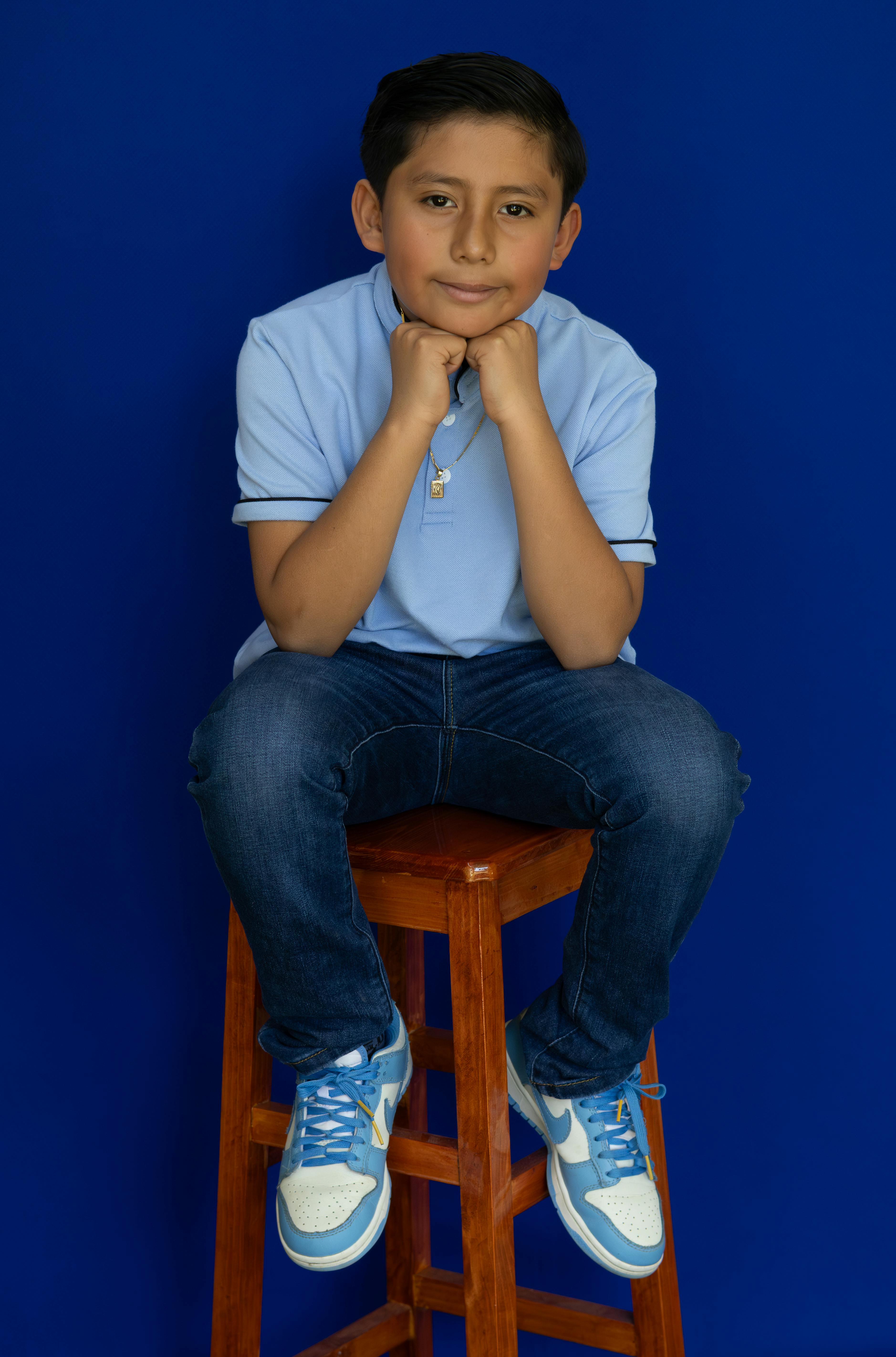 A young boy in casual blue attire seated on a wooden stool against blue backdrop.