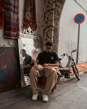 A young artisan sitting outdoors in Casablanca with tapestries and a mirror, exuding local charm.