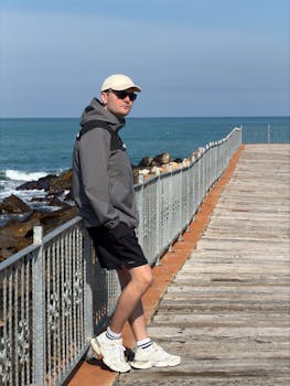 Casual man in jacket and cap standing on a pier by the ocean, enjoying a sunny day.