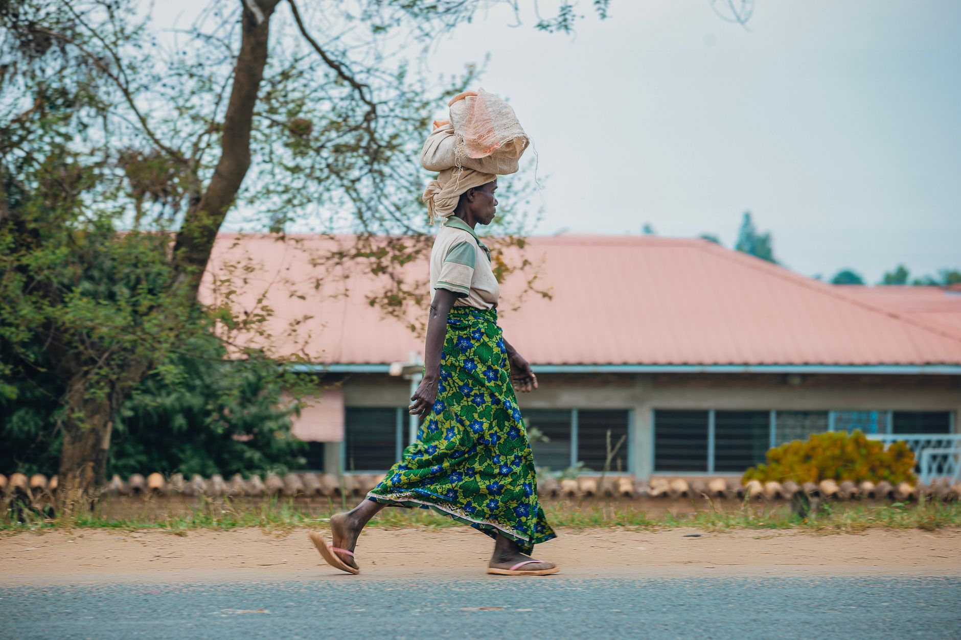 A woman in traditional attire walks outside carrying a basket on her head, showcasing cultural lifestyle.