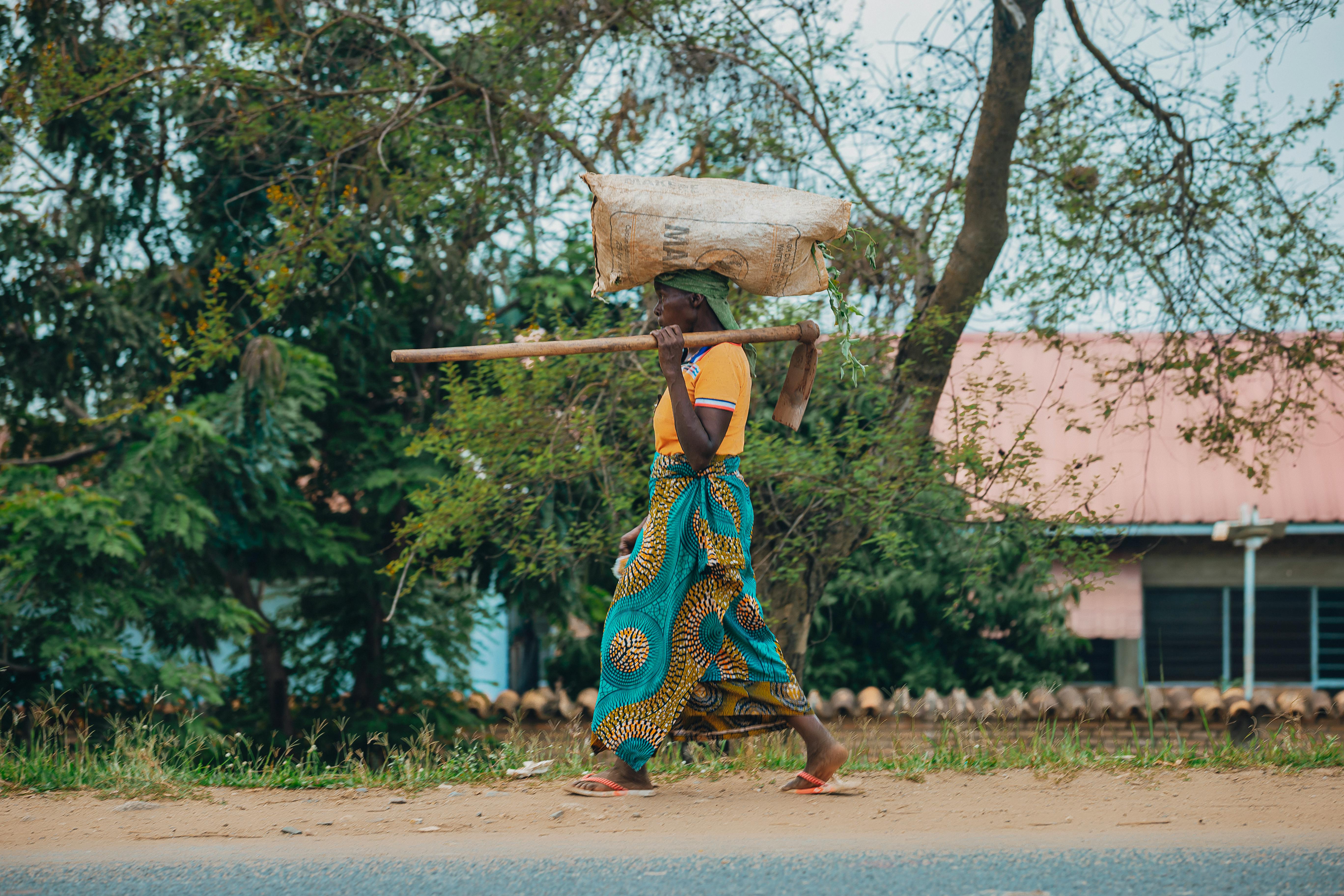 бесплатная Африканская женщина с мешком на голове идёт по сельской дороге под деревом. Стоковое фото
