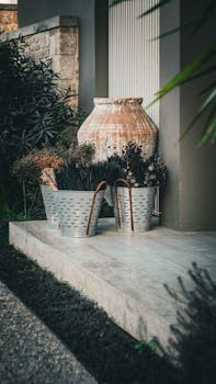 A rustic garden display featuring metallic buckets with dried plants and a large ceramic jar on steps.