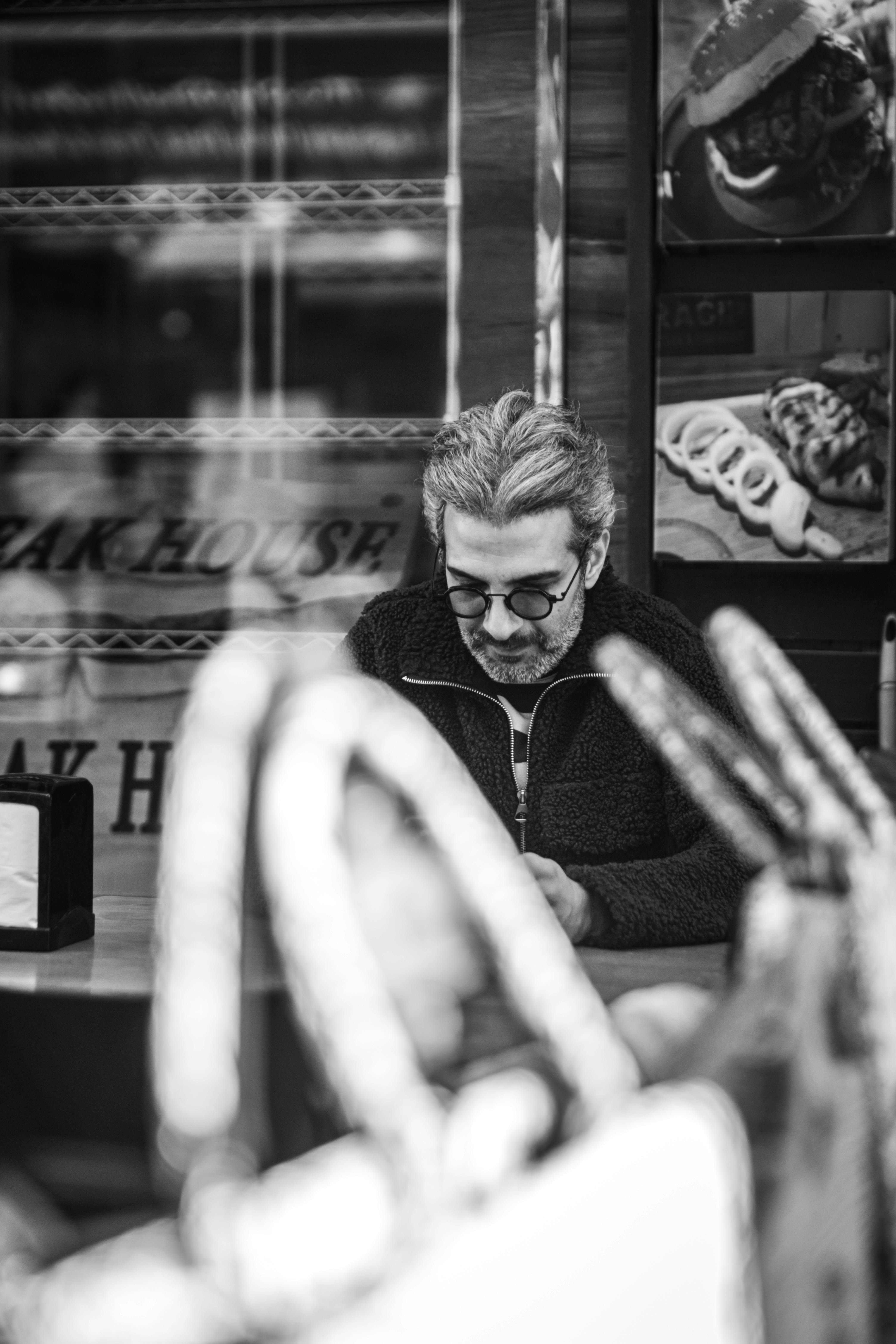 Free Thoughtful man with glasses sits alone in a cafe, captured in a monochrome street scene. Stock Photo