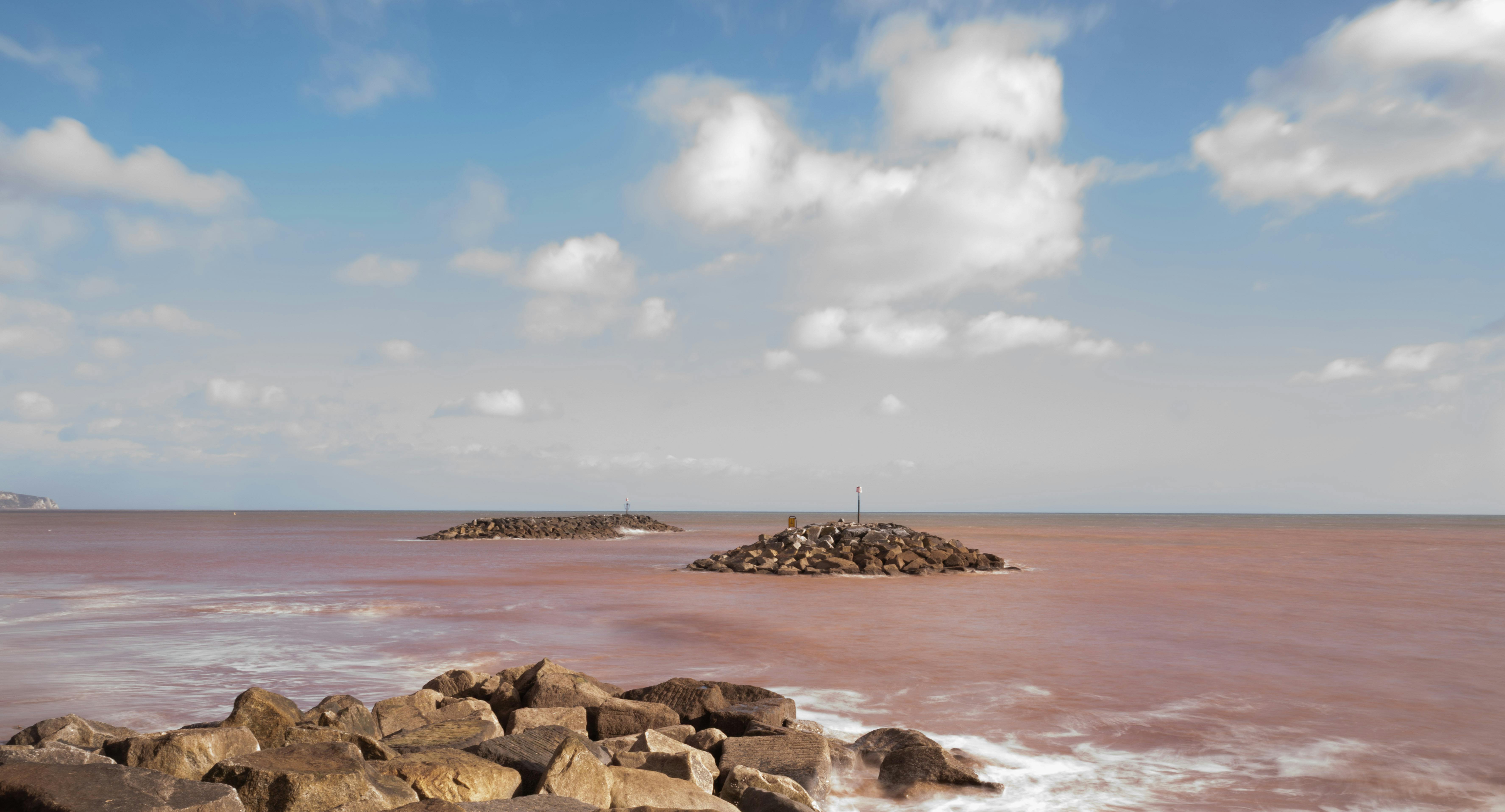 無料 ふわふわの雲が浮かぶ明るい青空の下、2 つの岩の多い防波堤が特徴的な静かな海の景色。 写真素材