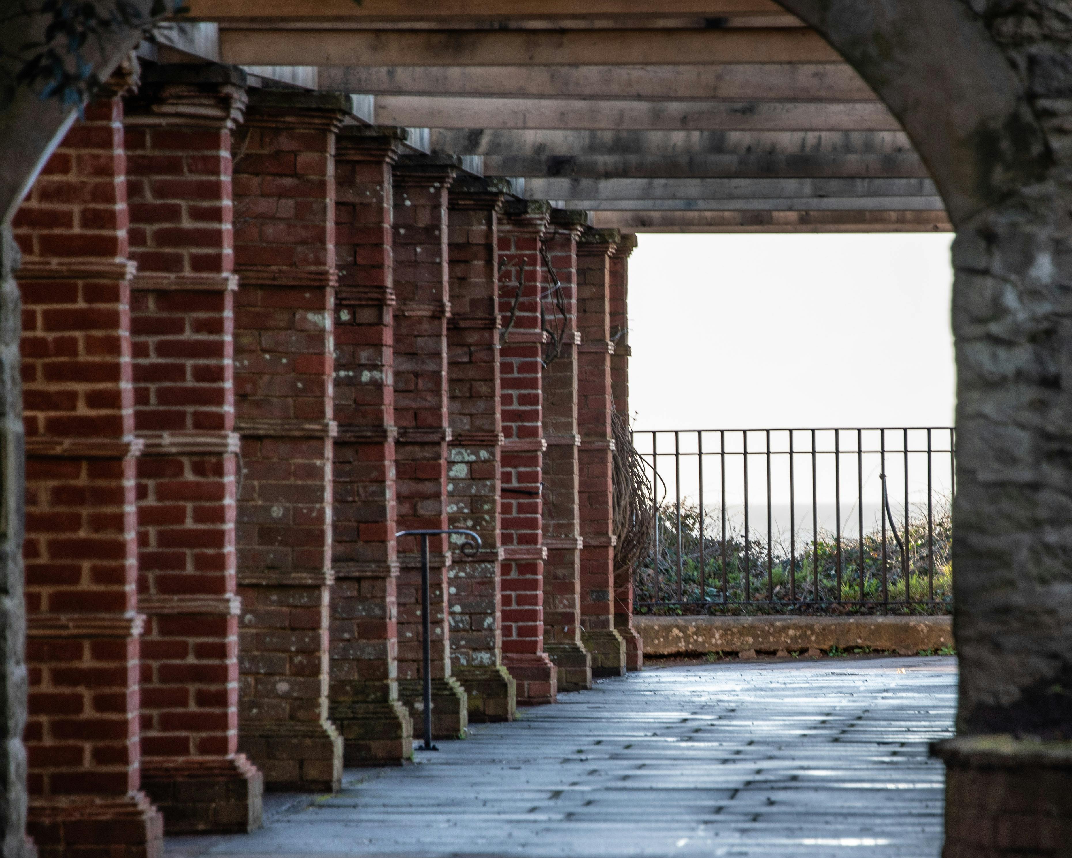 бесплатная Архитектурный снимок кирпичного колонного коридора, ведущего к железному перилу, с видом на улицу. Стоковое фото
