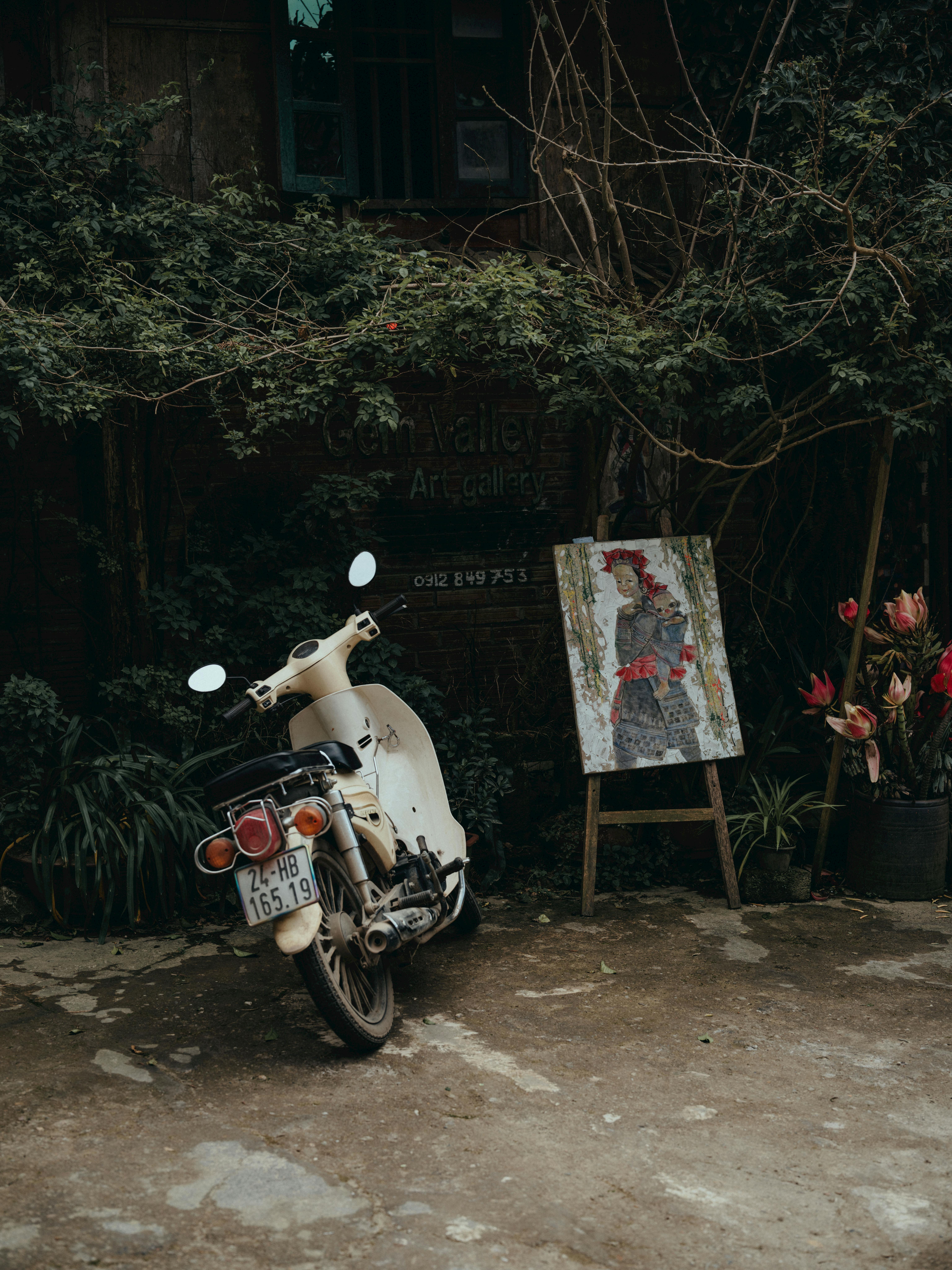 De franc Escúter retro aparcat al costat d'una galeria d'art enmig d'una vegetació exuberant a Lào Cai, Vietnam, que exhibeix pintura artística de paisatges. Foto d'estoc