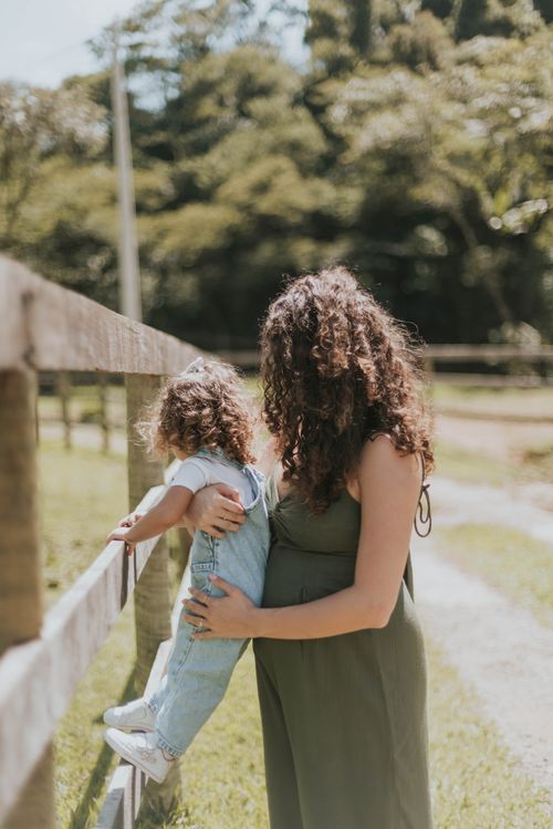 Free A woman holds a toddler by a wooden fence in a sunny outdoor setting, surrounded by trees. Stock Photo