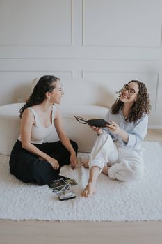 Two women sitting on a soft rug, sharing photos and enjoying laughter in a bright, cozy room.