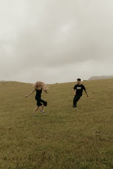 A couple enjoying a playful moment running across a grassy field under a cloudy sky.