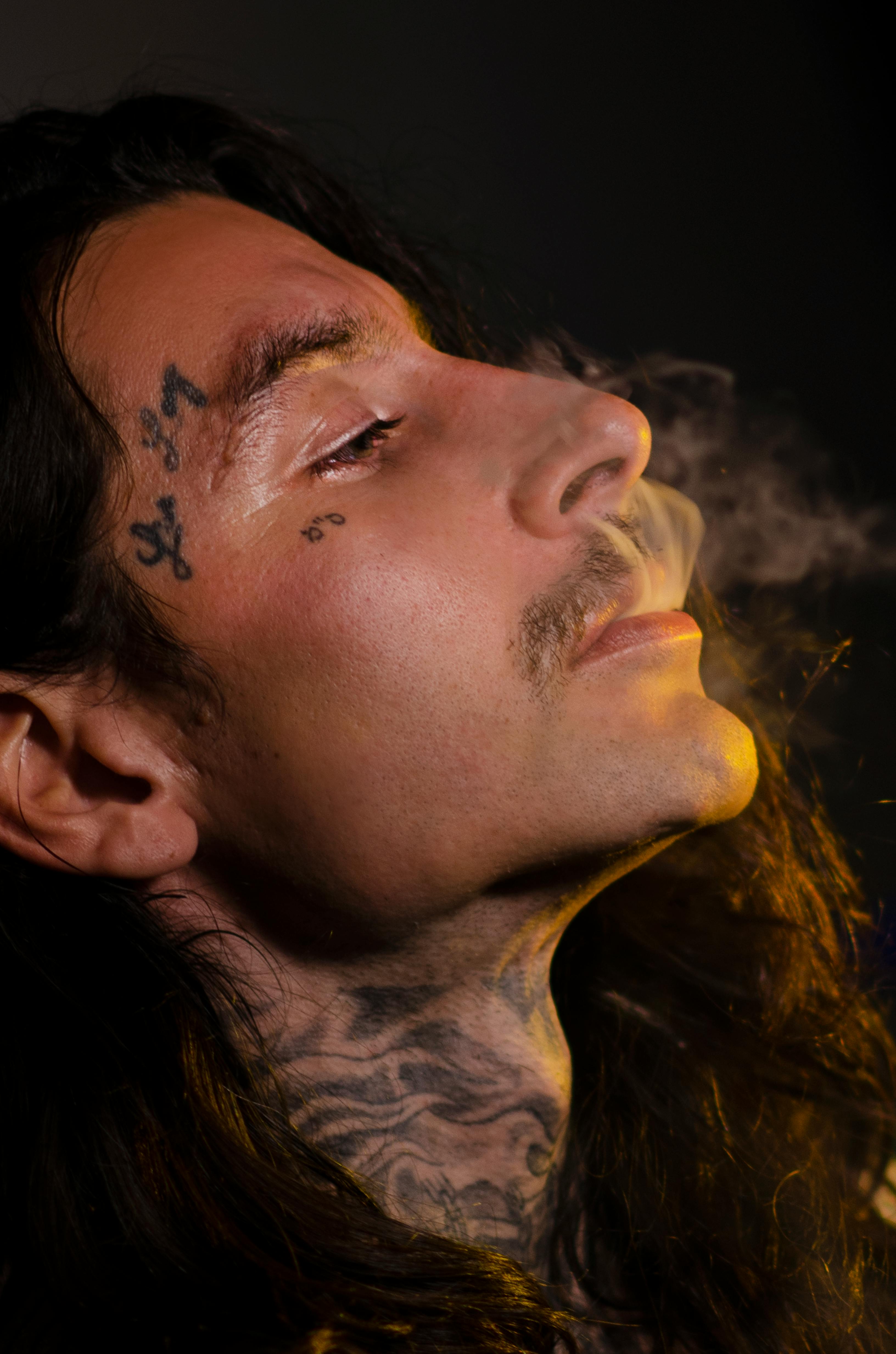 Free Close-up of a tattooed man exhaling smoke, highlighting facial tattoos and expressive mood. Stock Photo