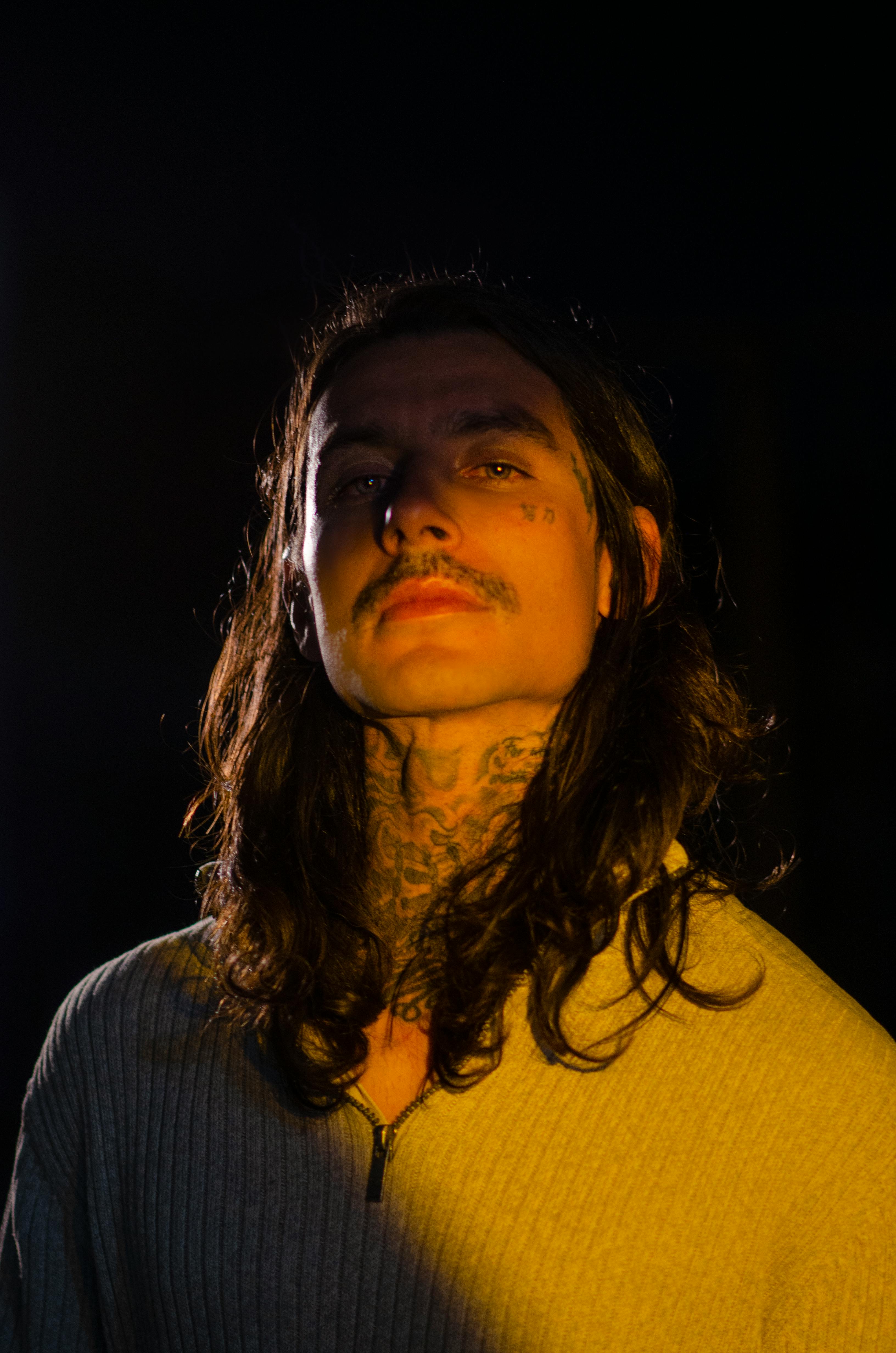 Gratuit Portrait saisissant d'un homme tatoué, aux cheveux longs et à la moustache, mis en valeur par une douce lumière dans l'obscurité. Photos