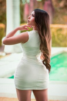 Stylish woman in a white dress with long hair posing by a pool on a sunny day.