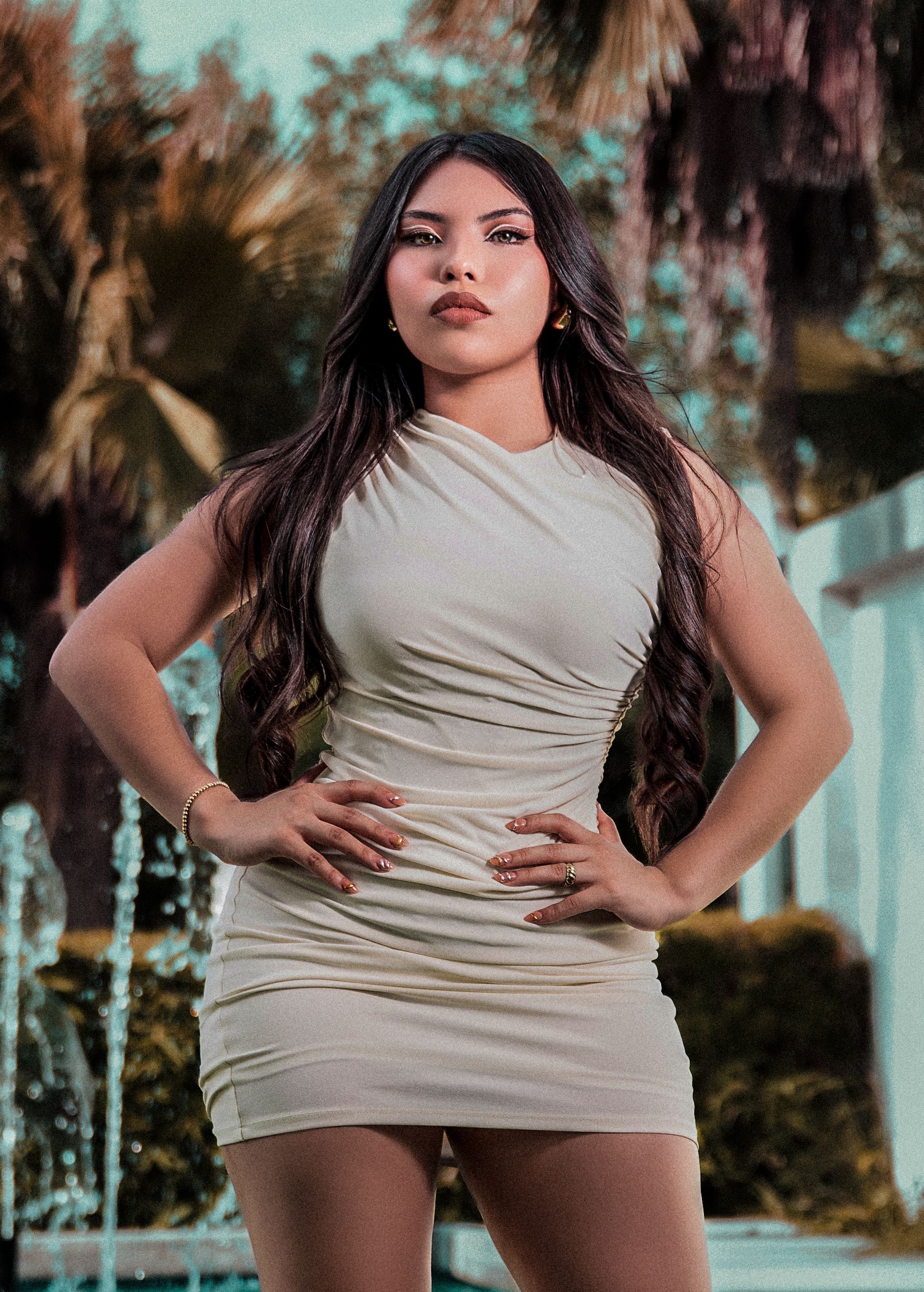 Confident woman in a stylish white dress poses in an elegant outdoor setting with palm trees.