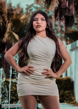 Confident woman in a stylish white dress poses in an elegant outdoor setting with palm trees.