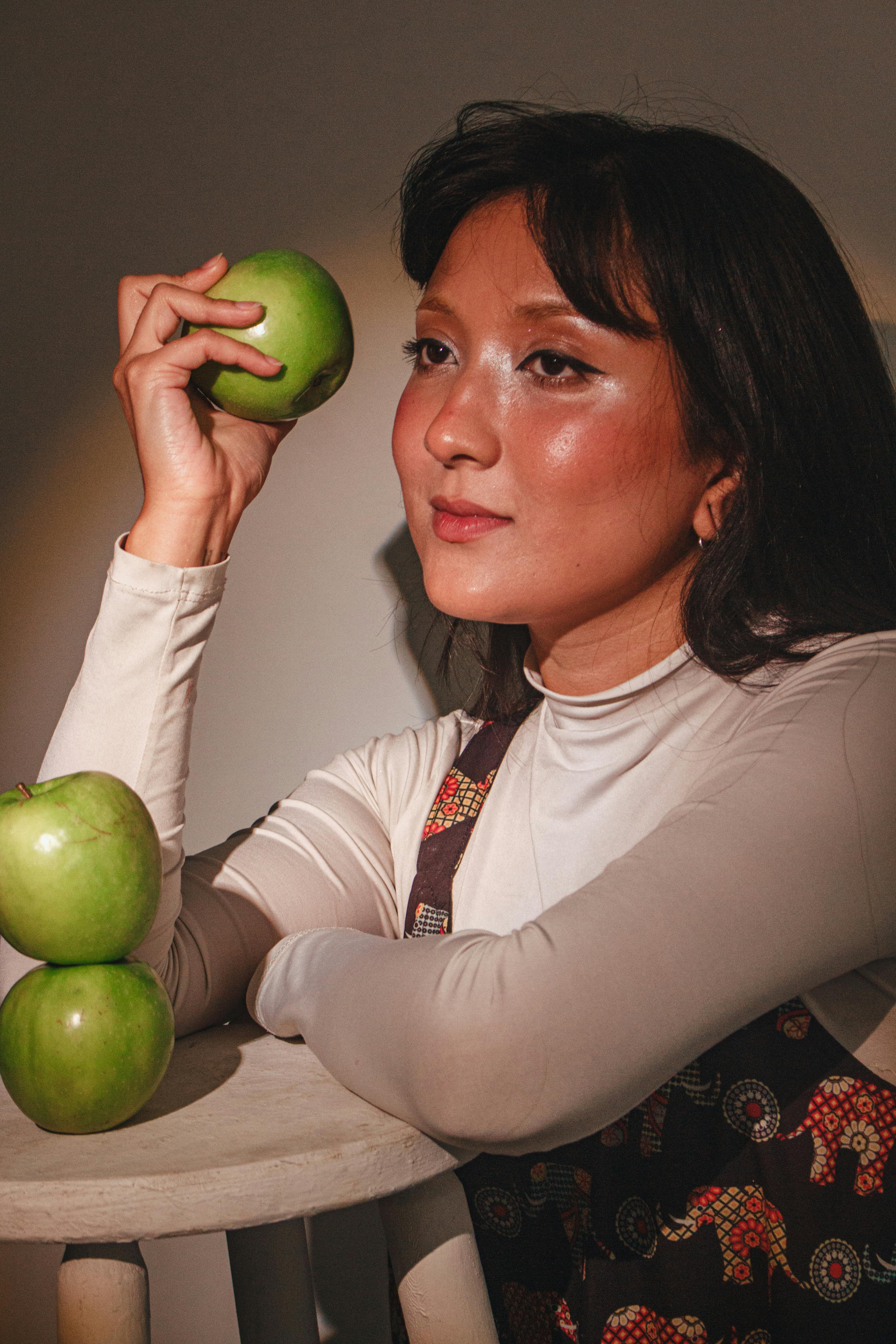 無料 暖かい照明の中で、女性が緑のリンゴを持ってポーズをとり、芸術的なポートレートを作り出しています。 写真素材